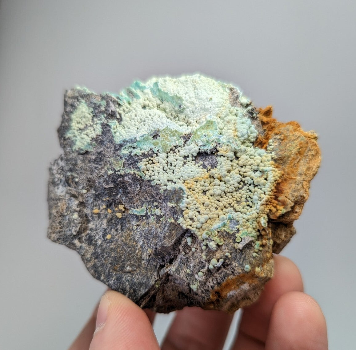 A rugged, irregularly shaped mineral specimen featuring a textured surface with patches of pale green and yellowish-white crystalline formations over dark gray rock.