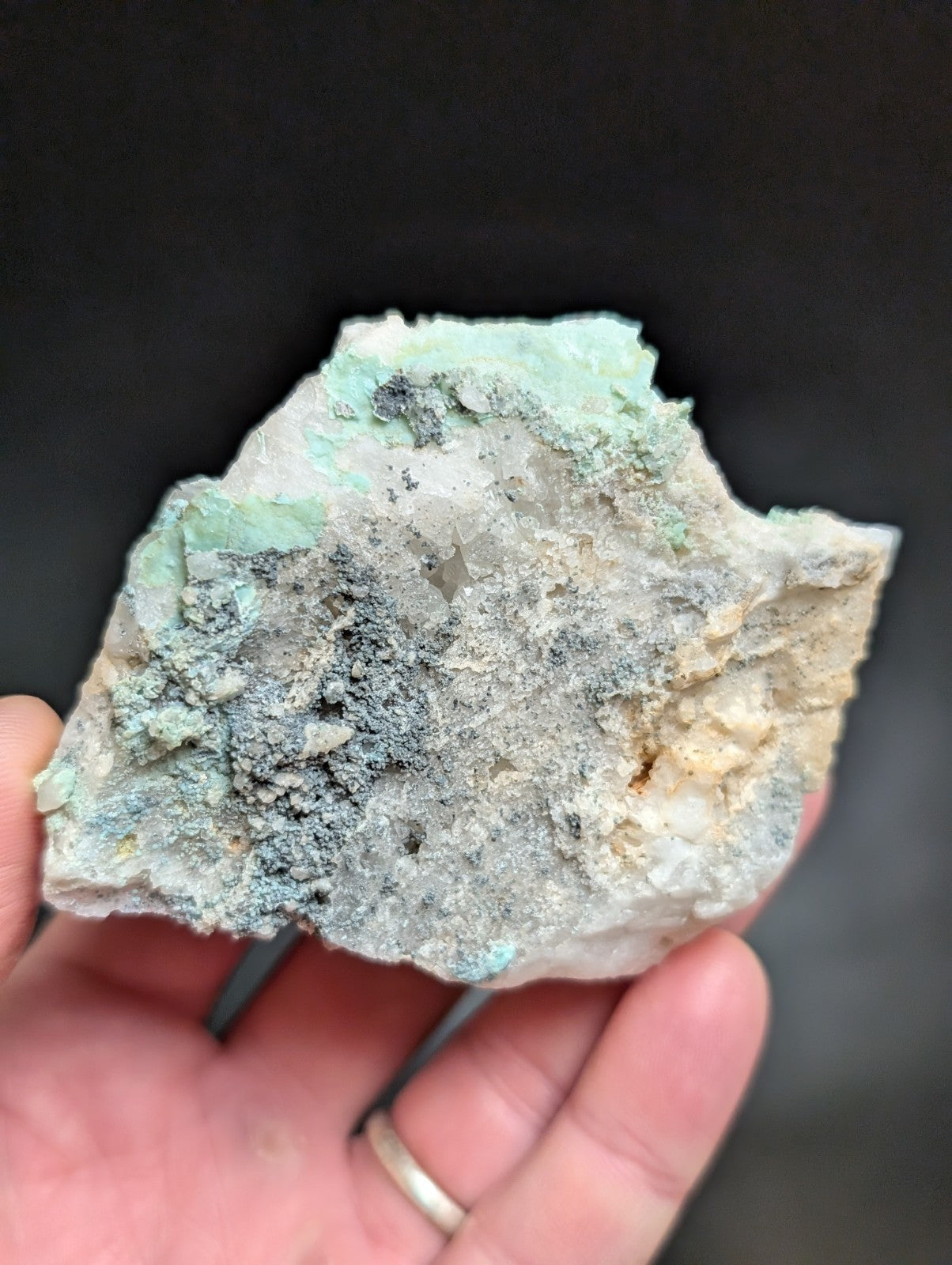 A hand holds a rough, irregularly shaped mineral specimen featuring patches of light green and grayish-blue patina over a lighter, off-white base.