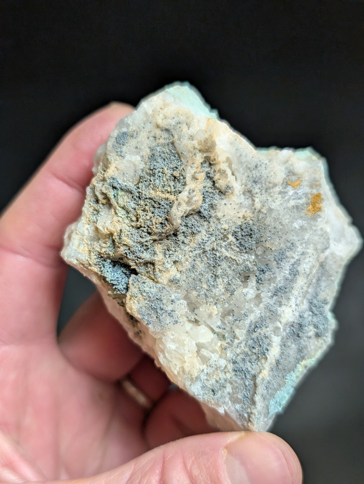 A hand holds a rough, irregularly shaped mineral specimen with a mottled surface of white, grayish-blue, and faint yellow patches.
