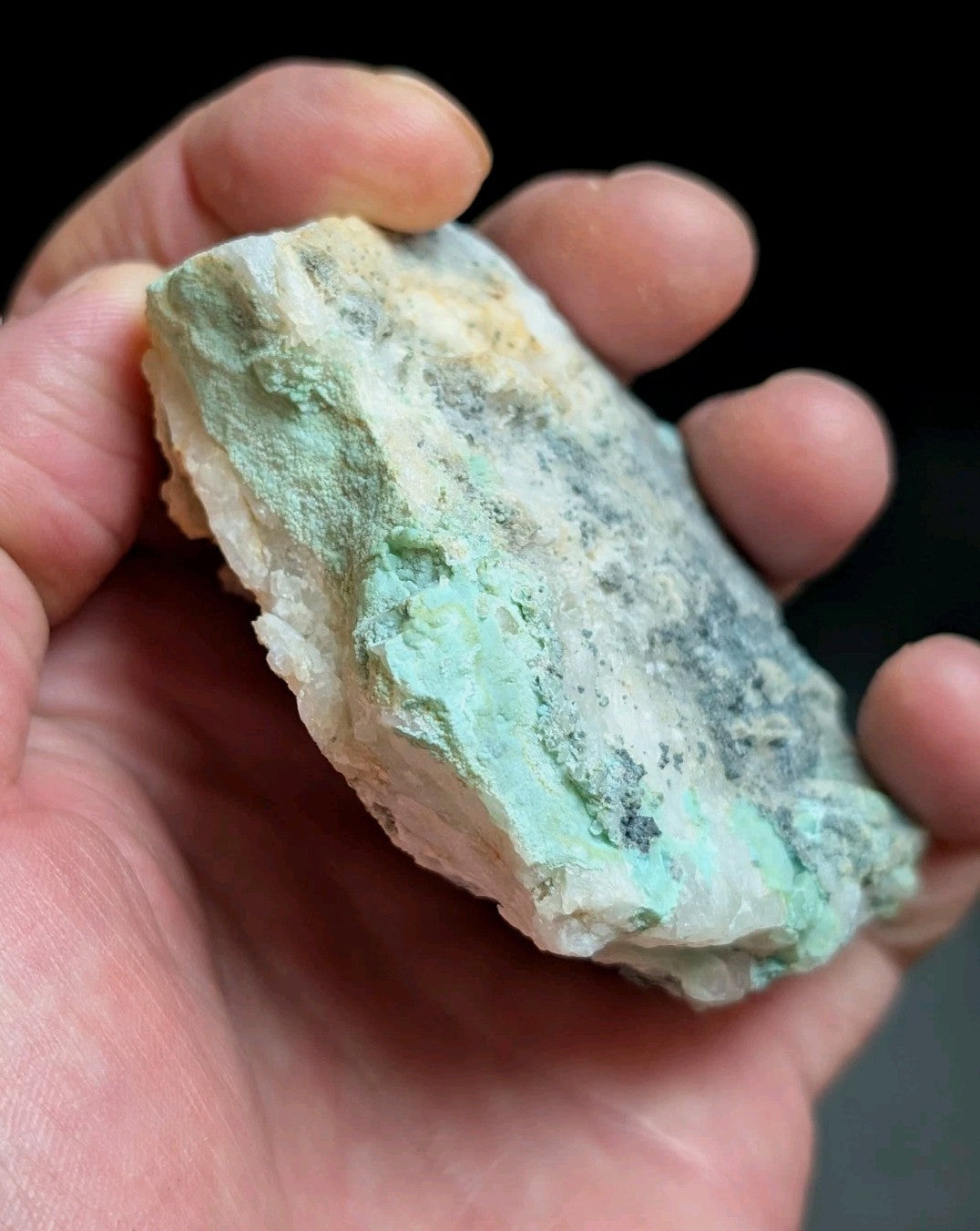 A hand holds a rough, irregularly shaped mineral specimen with patches of pale green and white, featuring subtle veining and natural textures.