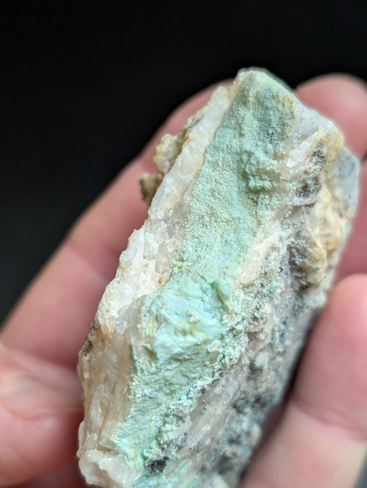A hand holds a rough, irregularly shaped mineral specimen with patches of pale green and white crystalline material.