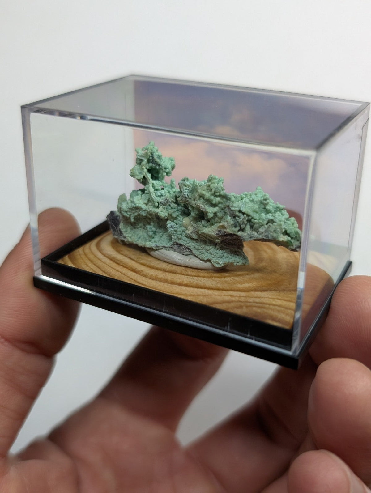 A miniature green copper mineral specimen displayed in a clear acrylic box with a wooden base.