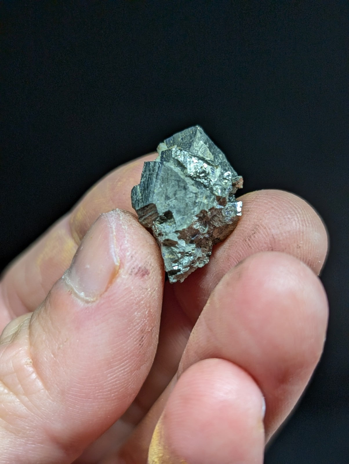 A metallic, faceted mineral specimen with a dark greenish-gray hue and reflective surfaces.