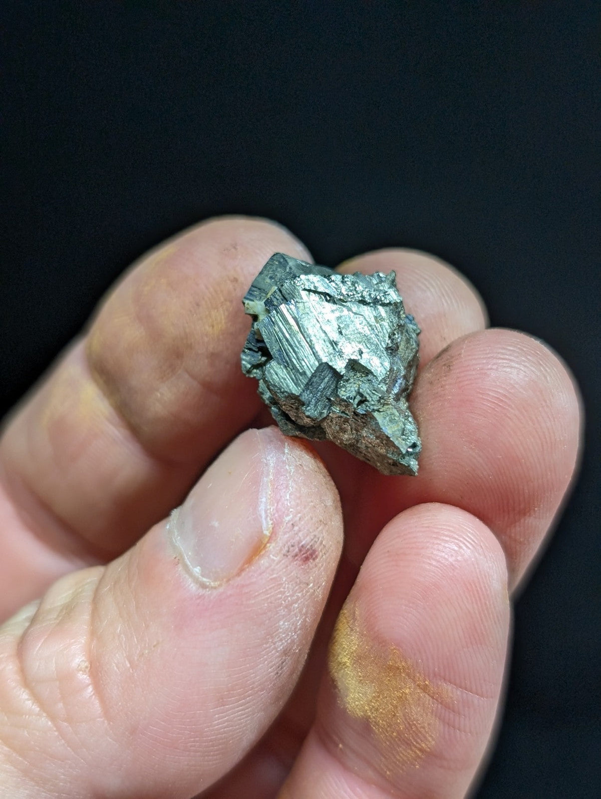 A small, irregularly shaped metallic mineral specimen with a greenish-silver sheen and visible crystalline facets.