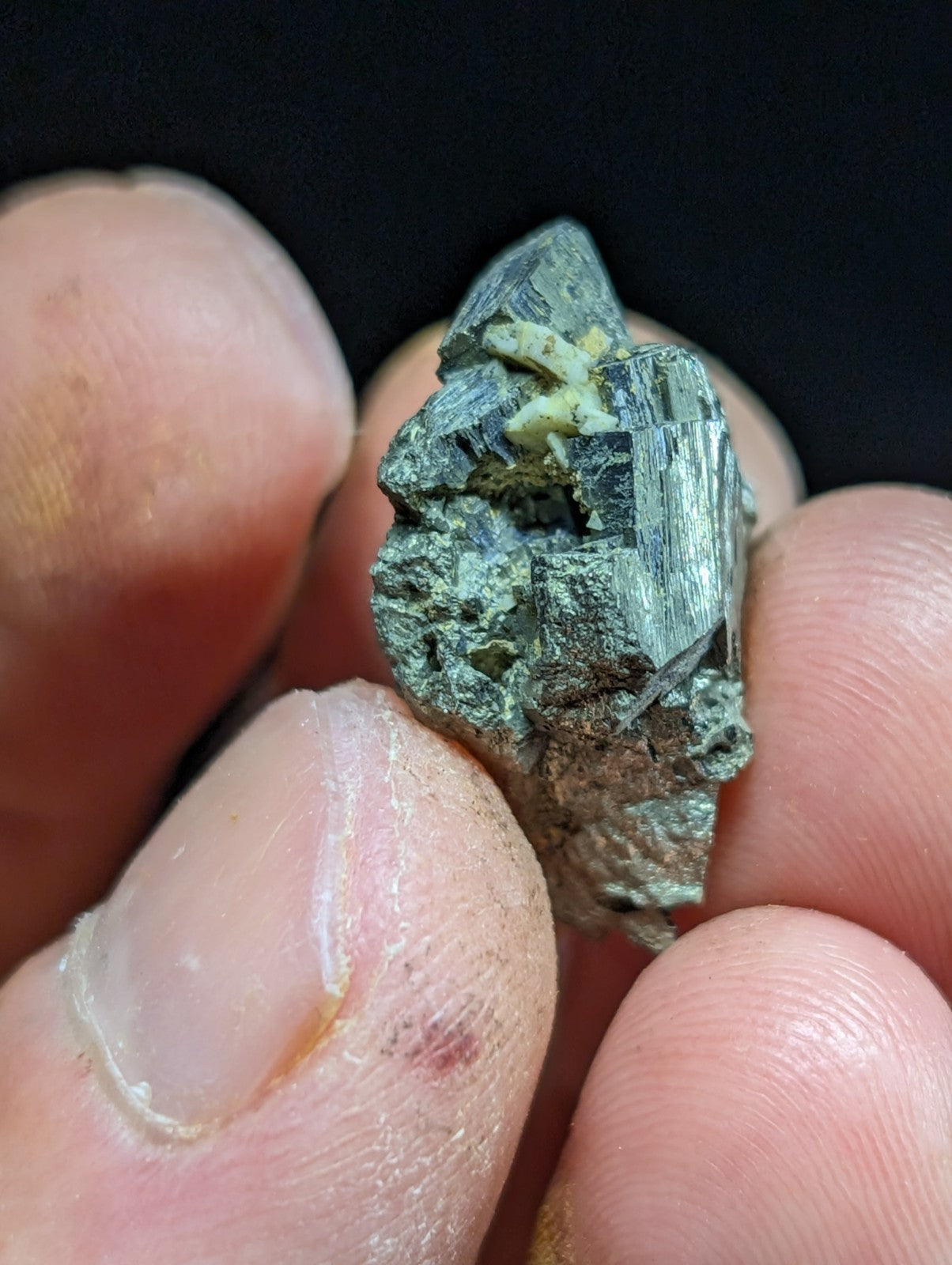 A rough, dark green mineral specimen with visible crystalline facets and a small patch of pale yellow material embedded within it.