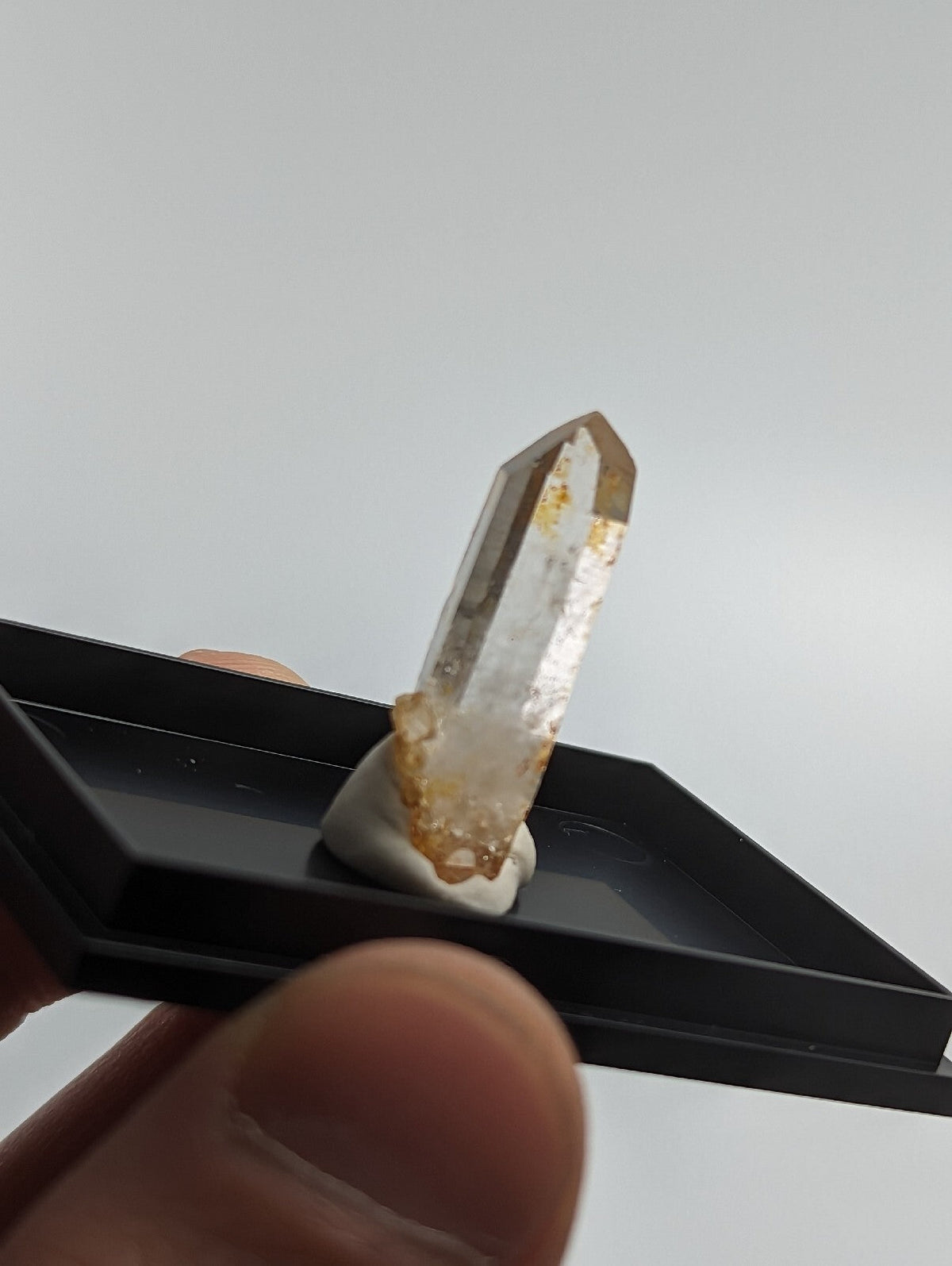A translucent, amber-tinted quartz crystal with a pointed tip and natural inclusions, mounted on a white base within a black display tray.