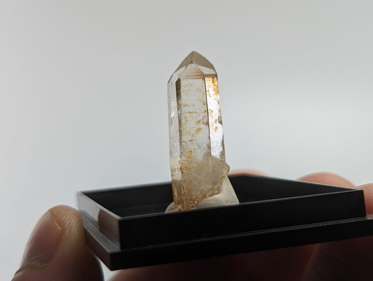 A translucent, golden-hued quartz crystal with a pointed tip and visible internal inclusions stands upright within a black display case.