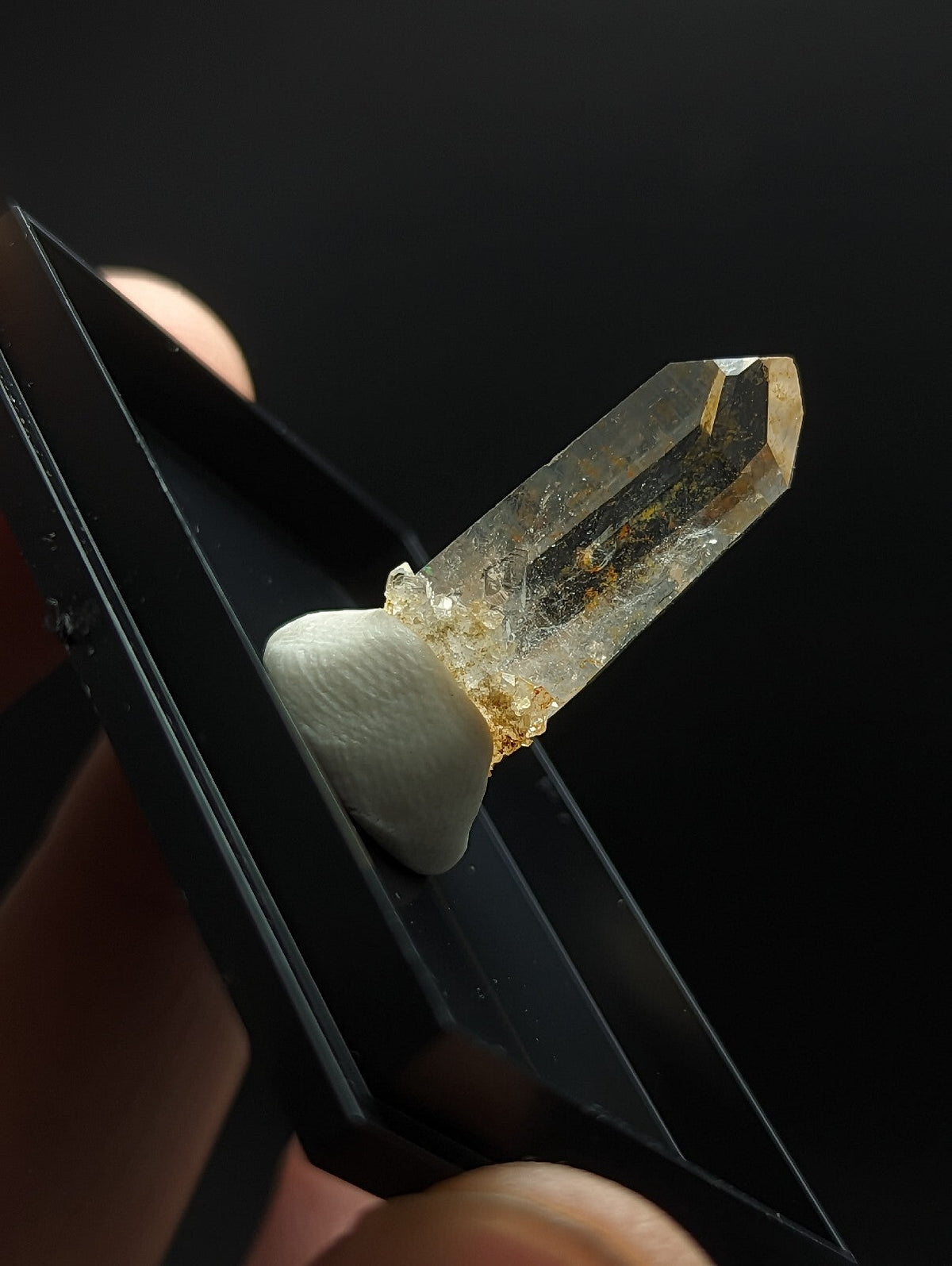 A translucent, golden-hued quartz crystal with a pointed tip and visible internal inclusions is held securely by black tweezers.