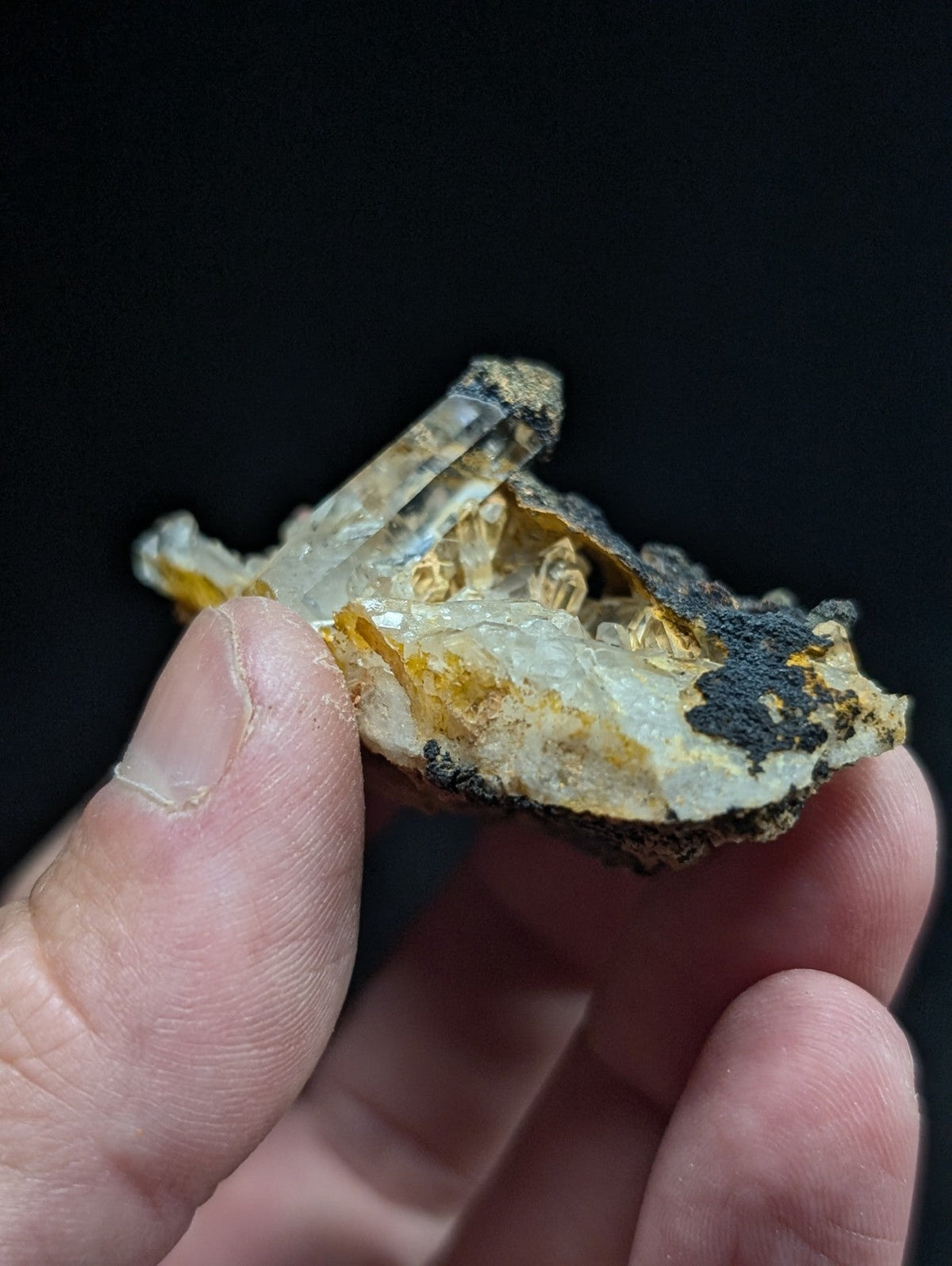 A translucent quartz crystal cluster with golden-yellow and dark mineral inclusions, held gently between fingers.