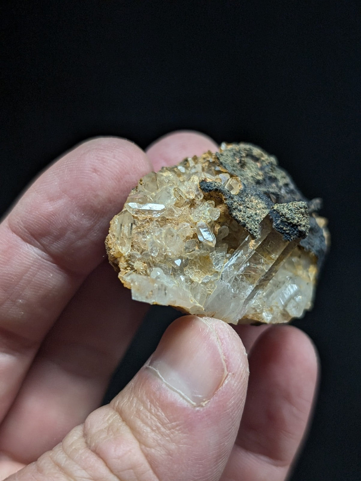 A hand holds a rough, translucent quartz crystal specimen with golden-brown matrix and dark mineral inclusions.