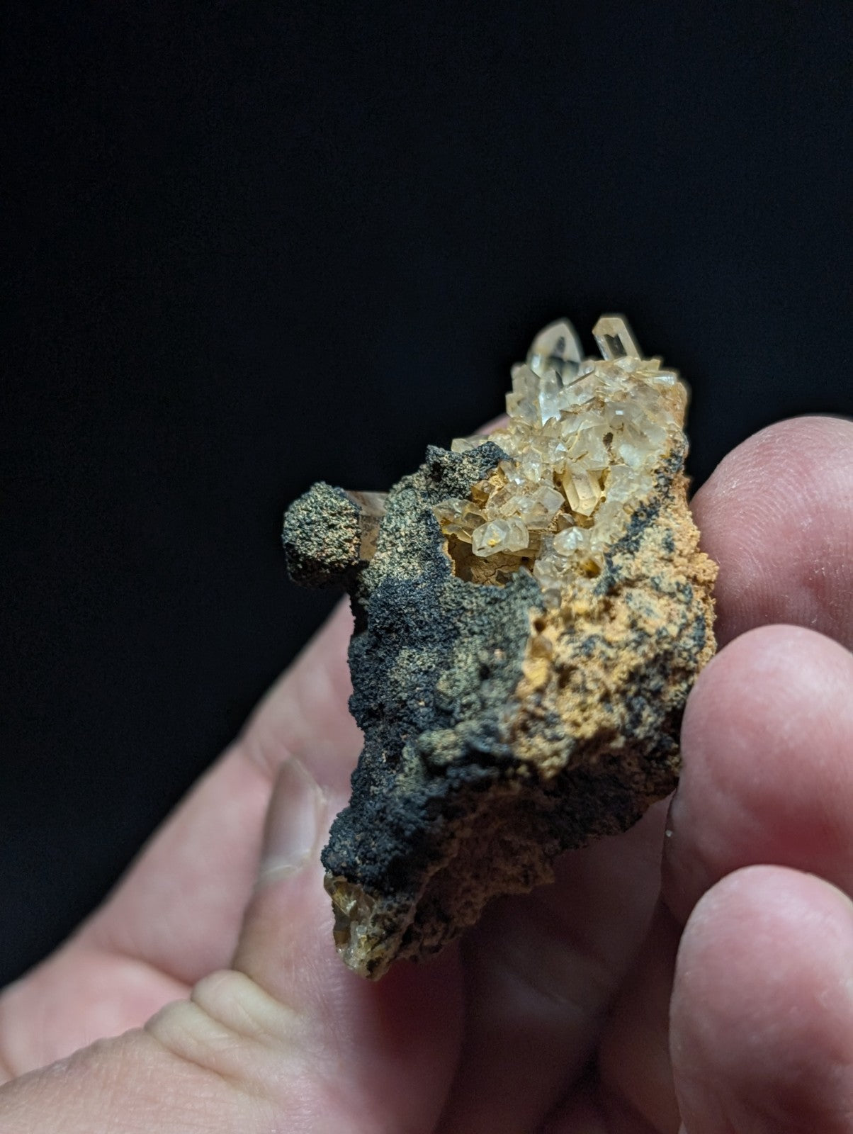 A hand holds a mineral specimen featuring translucent, crystalline formations atop a rugged matrix of dark and orange-brown rock.
