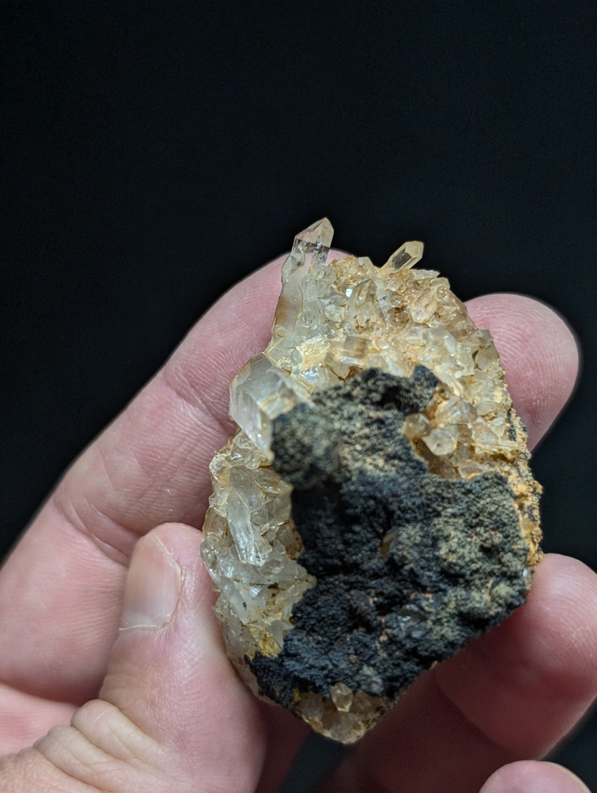 A hand holds a natural quartz crystal cluster with translucent, pale yellowish crystals growing from a dark gray matrix.