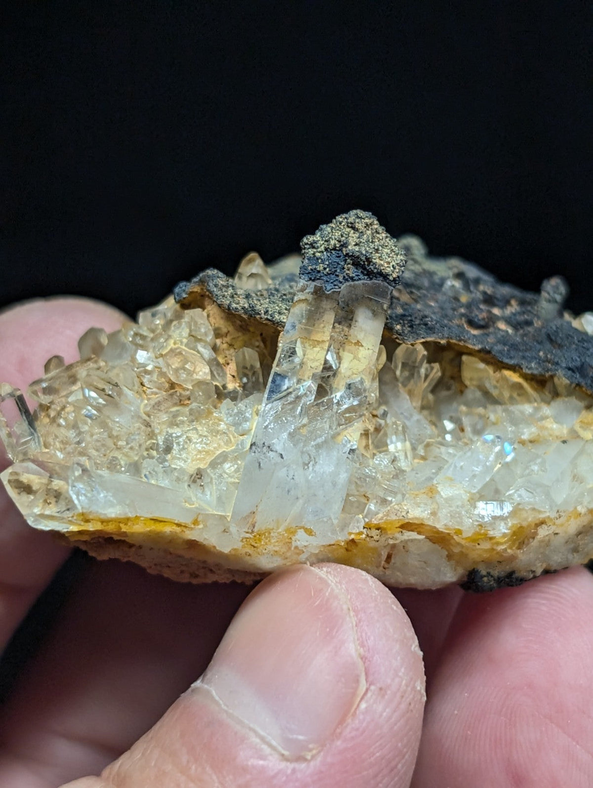 A translucent quartz crystal cluster with clear, faceted points emerging from a rough, earthy matrix of yellow and dark mineralized rock.