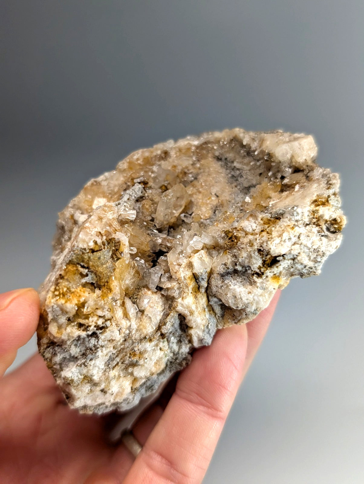 A hand holds a quartz crystal cluster featuring translucent solution quartz and cookeite in gray and yellow matrix