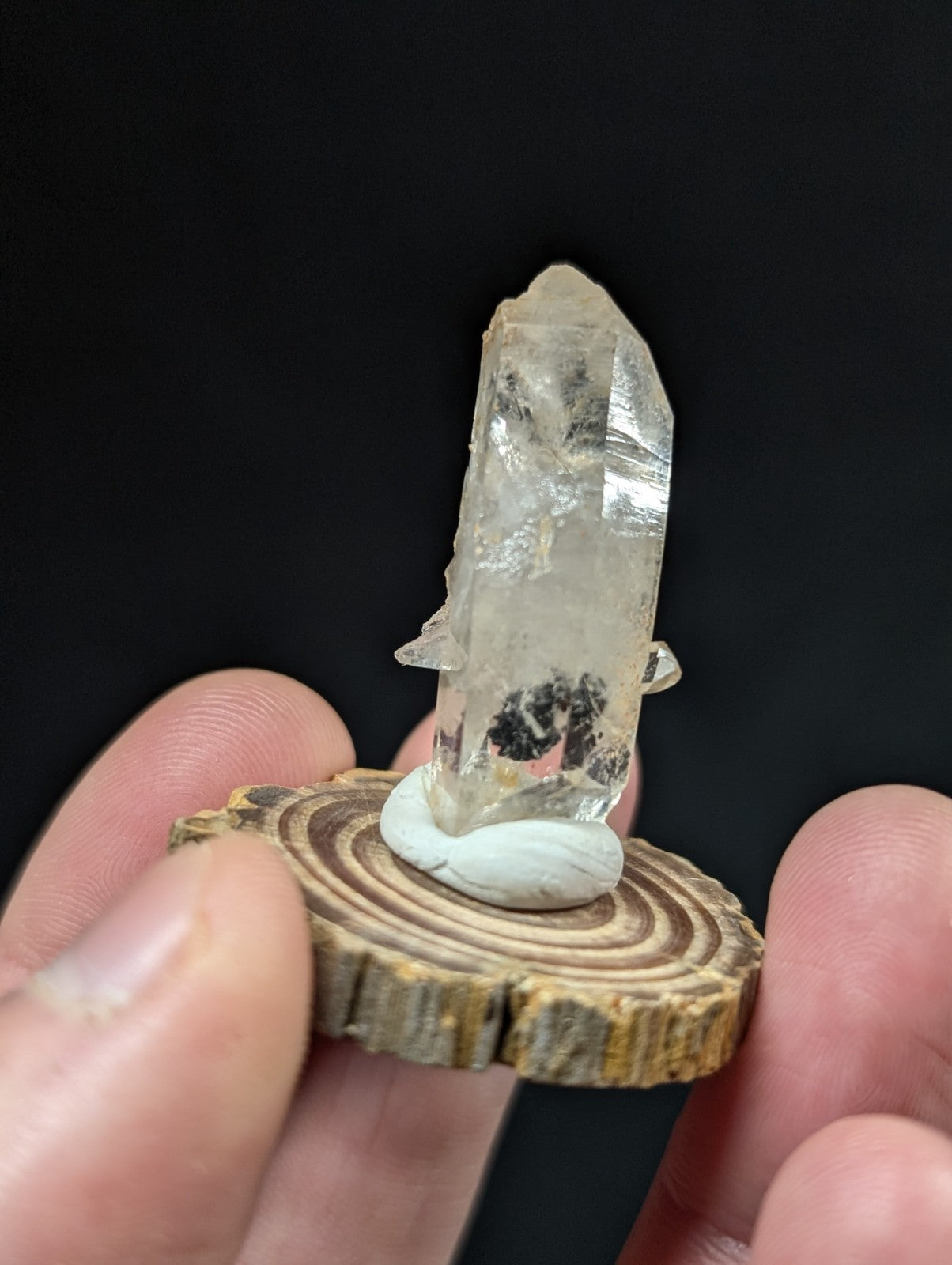A clear quartz crystal with natural inclusions, mounted on a white marble base and displayed atop a rustic wooden slice.