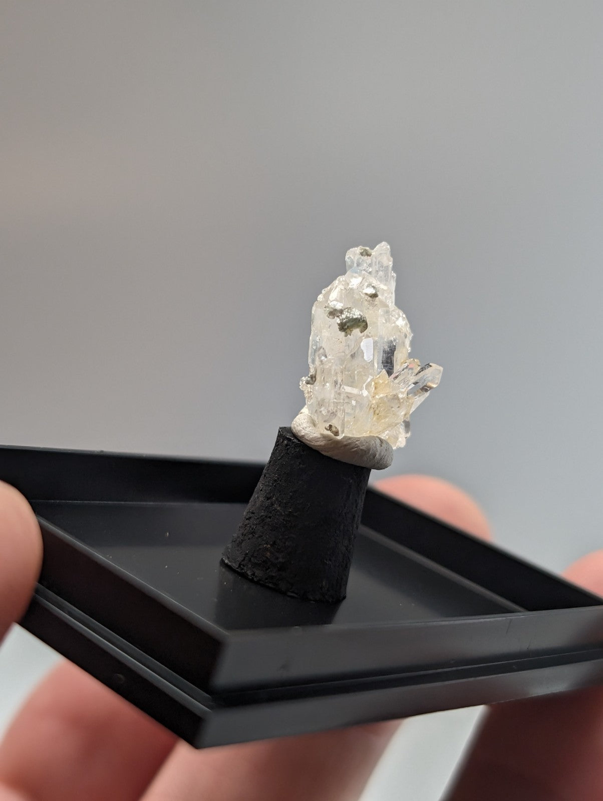 A translucent, crystalline mineral specimen with embedded metallic flecks sits atop a black cylindrical base within a sleek black display tray.