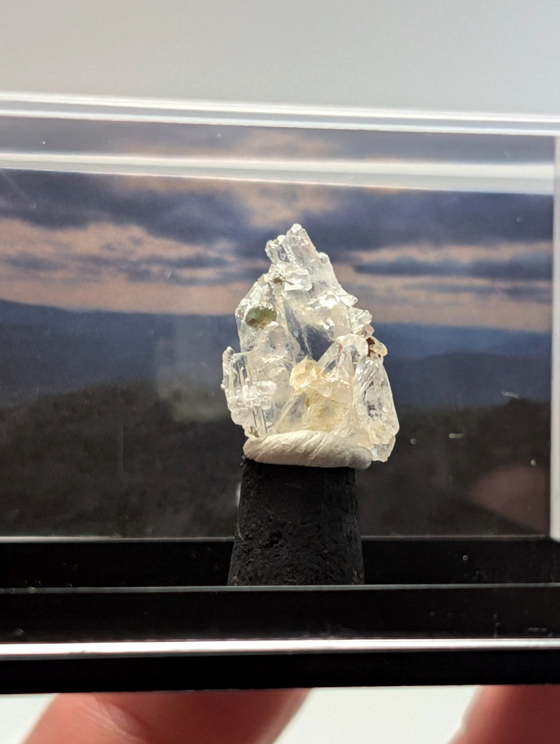 A translucent, crystalline mineral specimen with jagged edges and a pale yellowish tint, displayed on a black pedestal within a clear protective case.
