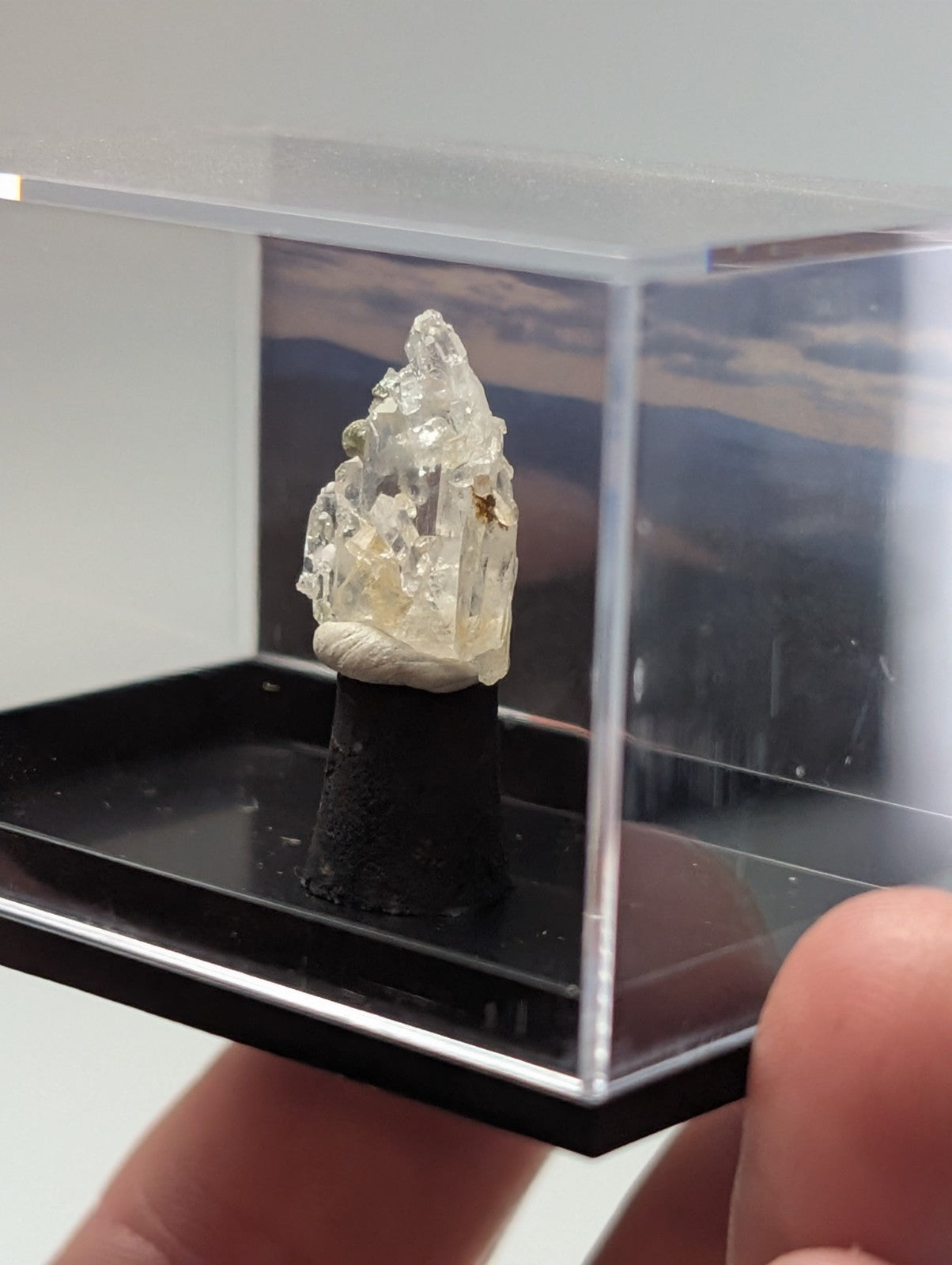 A translucent, crystalline mineral specimen with a rough, jagged surface and subtle yellowish tint is displayed on a black pedestal inside a clear acrylic display case.