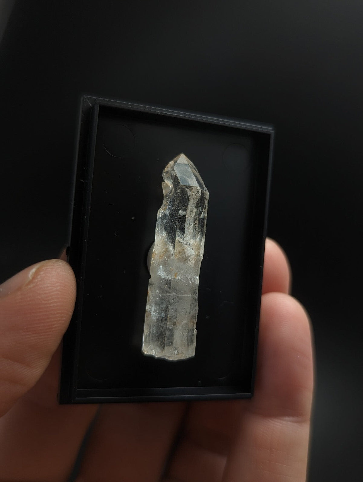 A clear quartz crystal with a pointed tip and natural inclusions, displayed inside a black rectangular box.