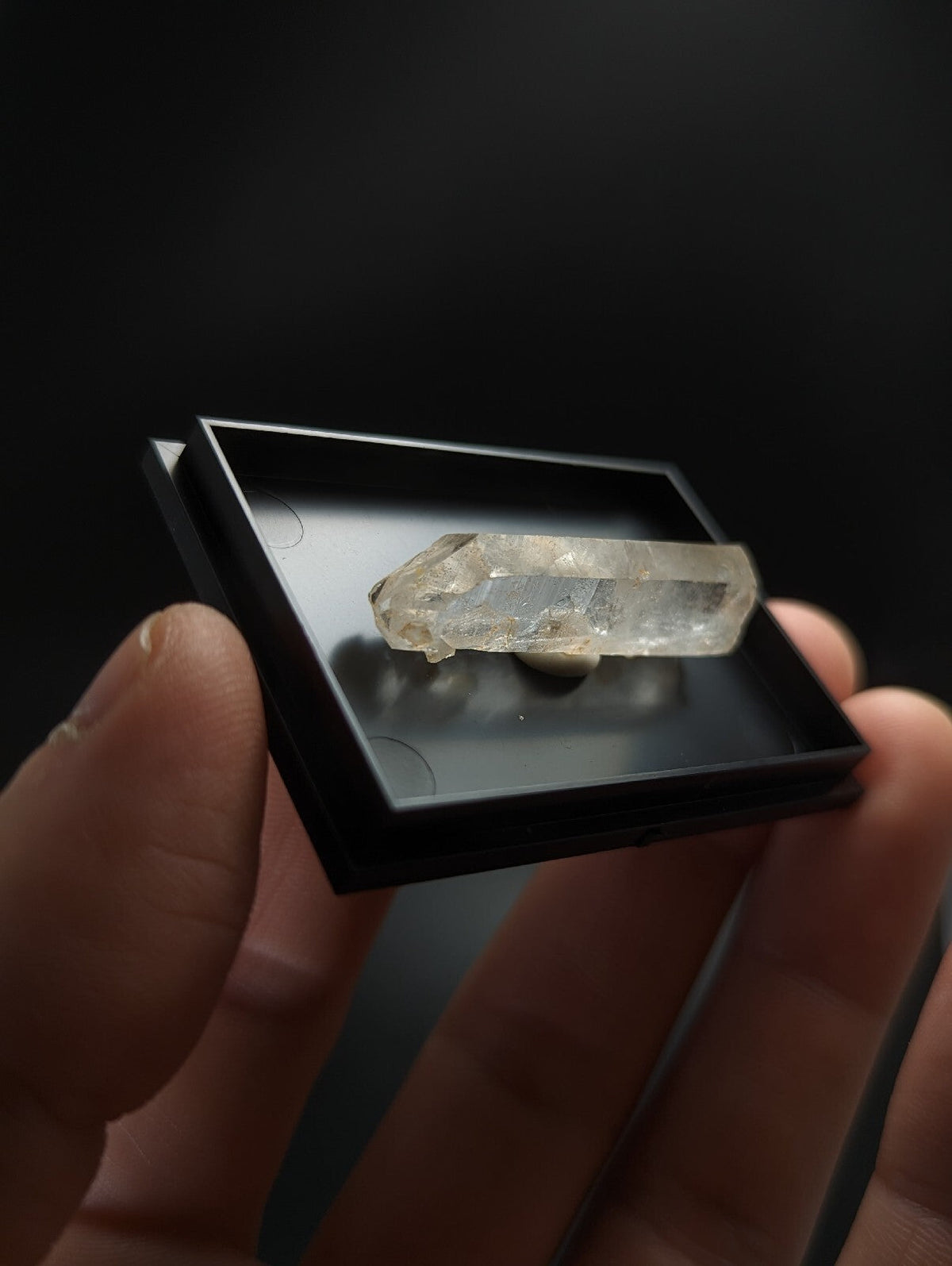 A translucent, clear quartz crystal with natural facets and subtle internal inclusions rests inside a sleek black display box.
