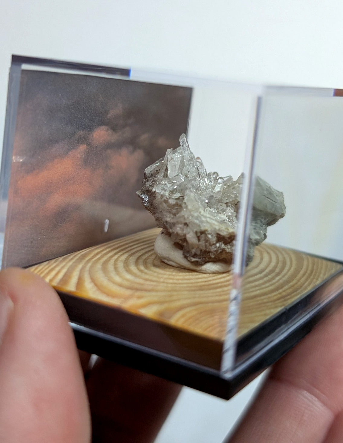 A translucent, crystalline mineral specimen with a metallic sheen rests on a wooden base inside a clear acrylic display case.