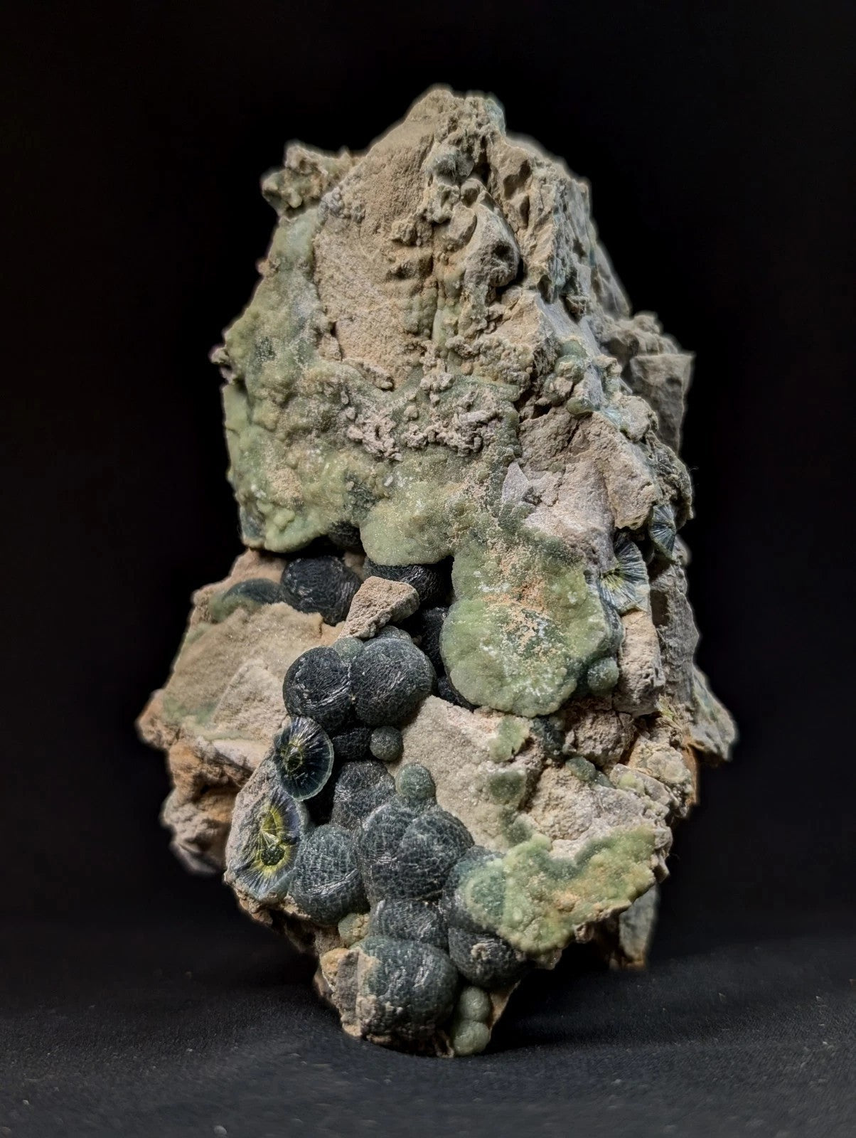 A rugged, irregularly shaped rock specimen featuring a cluster of dark green to black spherical formations nestled within lighter beige and grayish matrix material.