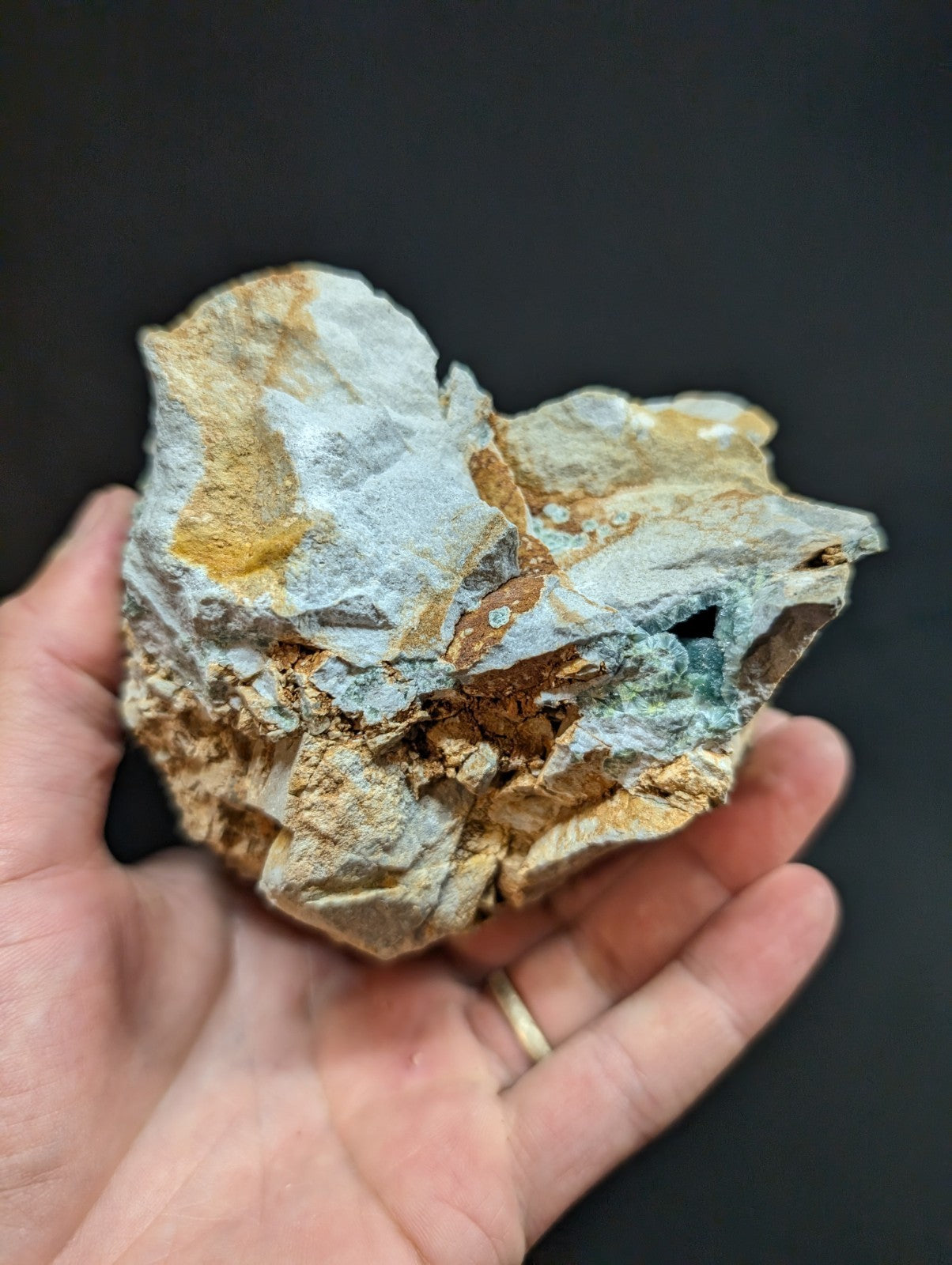 A hand holds a rough, irregularly shaped mineral specimen with patches of pale gray, golden yellow, and hints of greenish-blue veining.