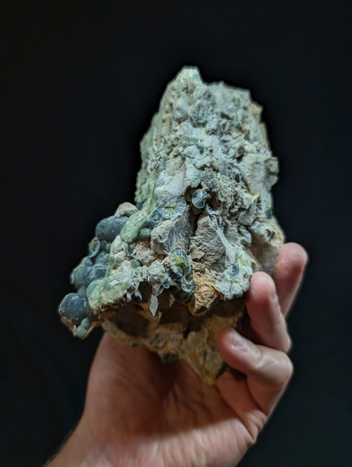 A hand holds a rough, irregularly shaped mineral specimen with patches of greenish-blue and gray hues, featuring embedded rounded nodules.