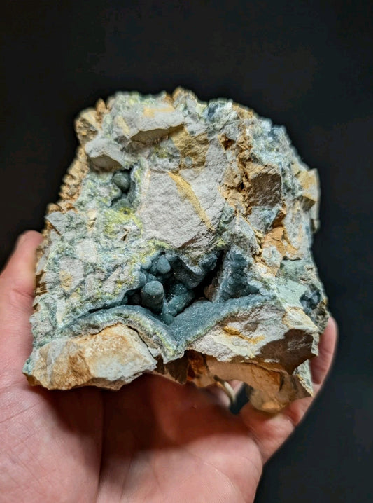 A hand holds a rugged, irregularly shaped mineral specimen featuring a striking contrast between its pale gray and yellowish rock matrix and the deep blue-green spherical crystals nestled within.