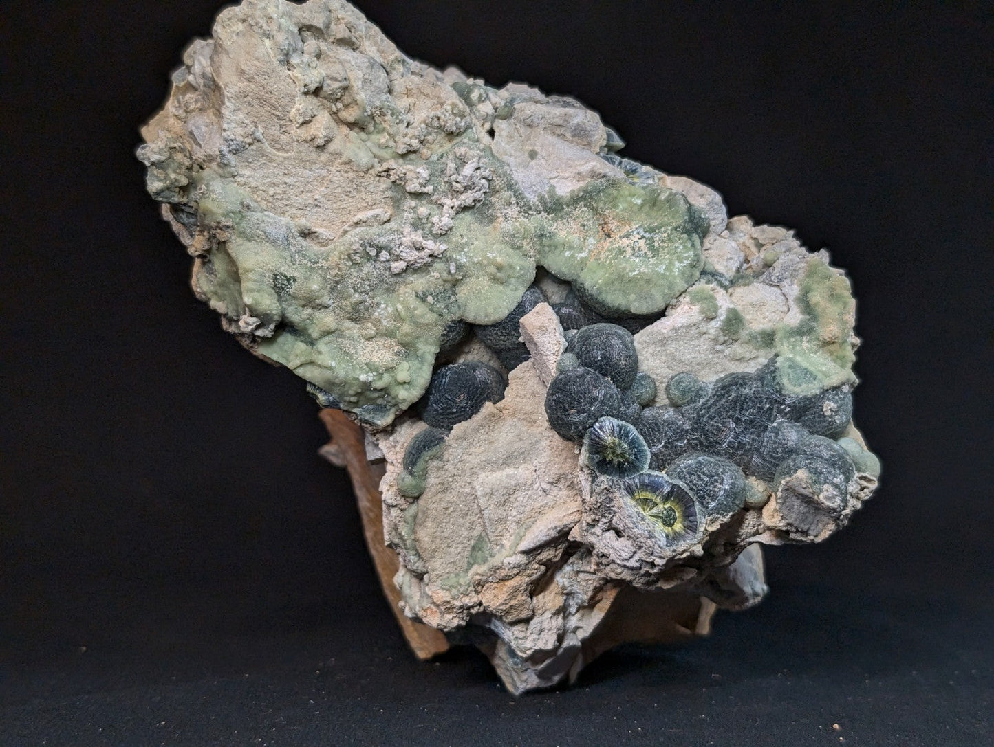 A rugged, naturally formed mineral specimen featuring a textured matrix of light green and grayish hues with clusters of dark, spherical formations.