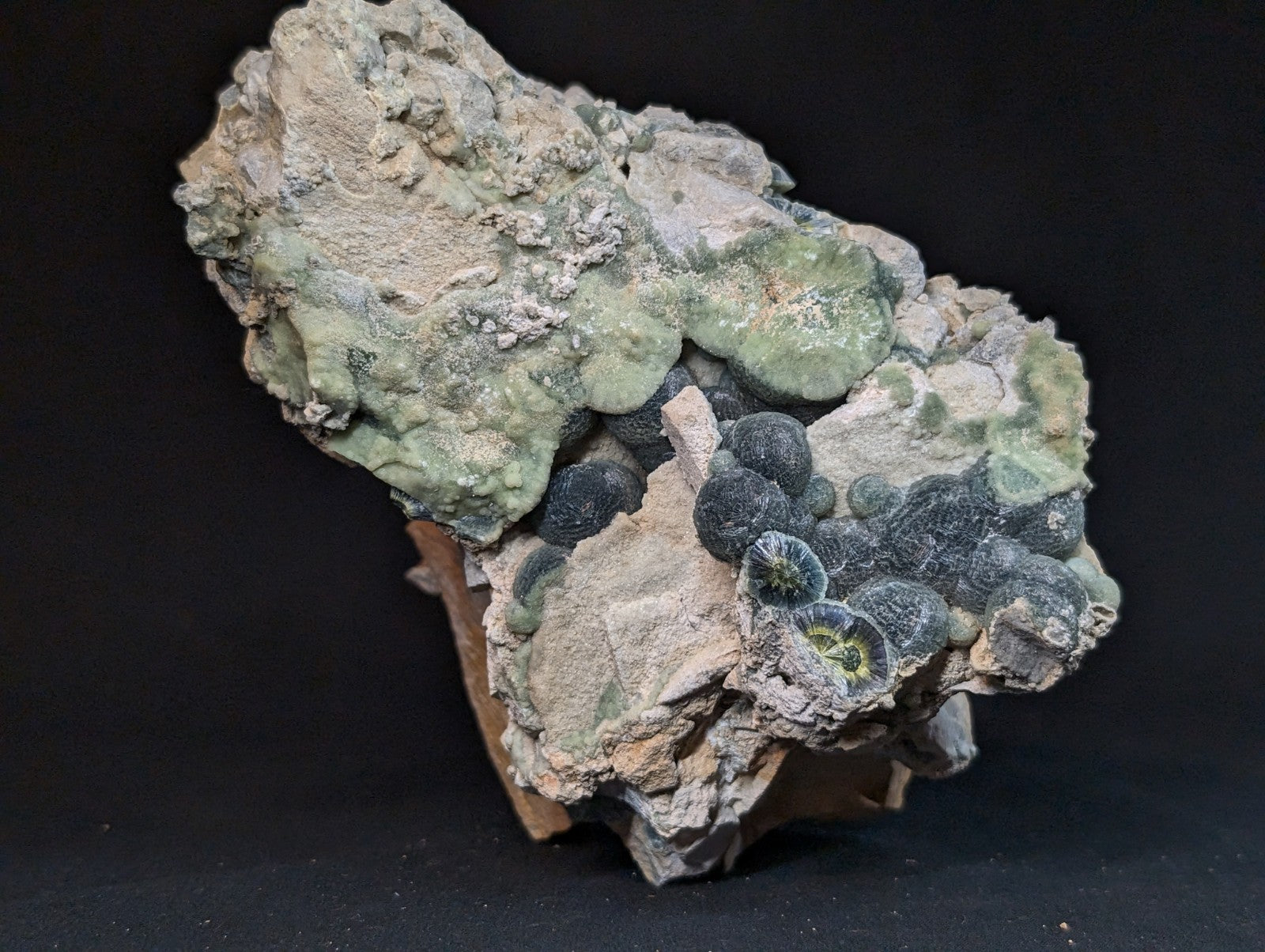 A rugged, naturally formed mineral specimen featuring a textured matrix of light green and grayish hues with clusters of dark, spherical formations.