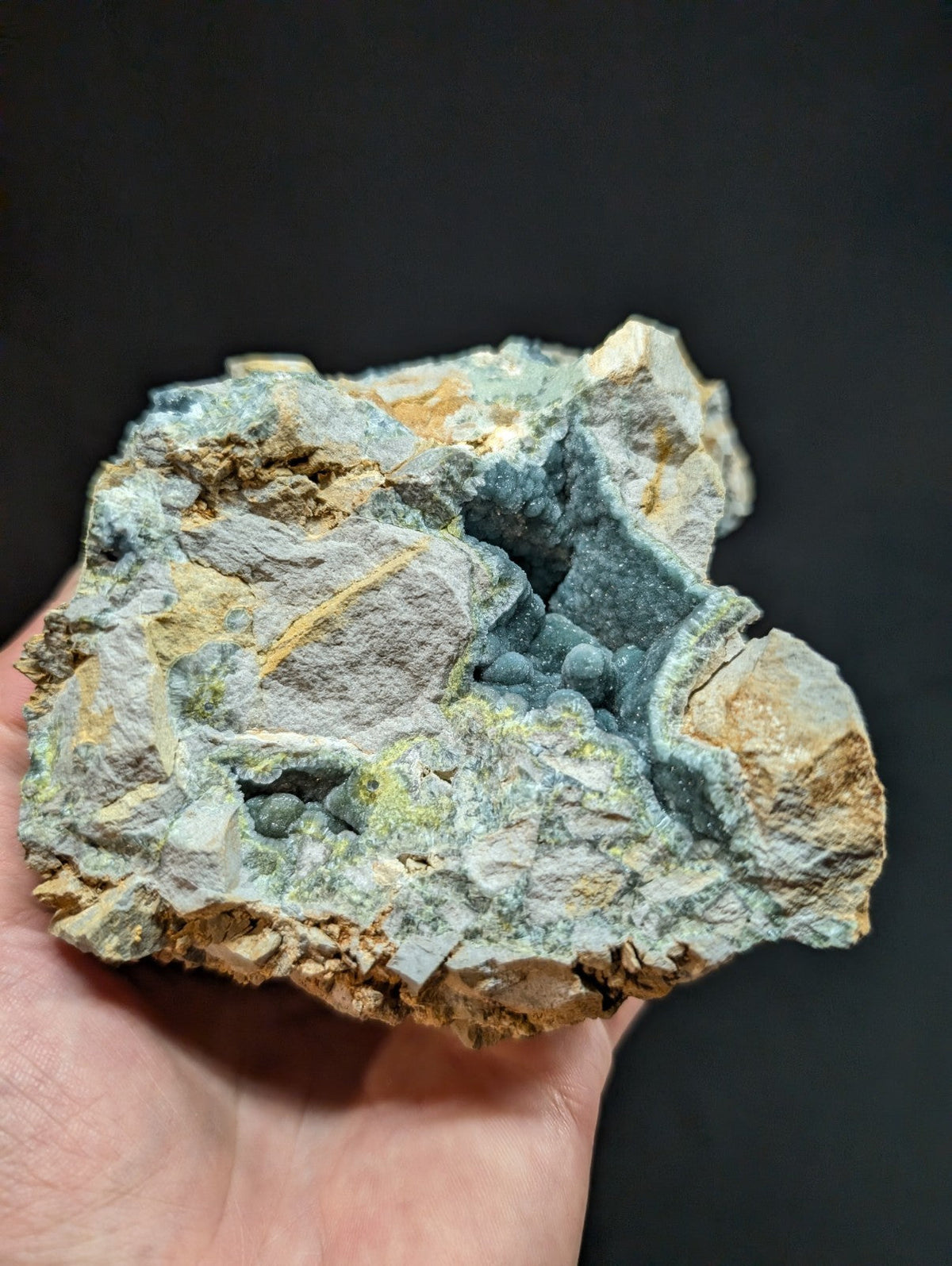 A hand holds a rough, irregularly shaped mineral specimen featuring patches of blue-green crystals embedded in gray and tan rock.