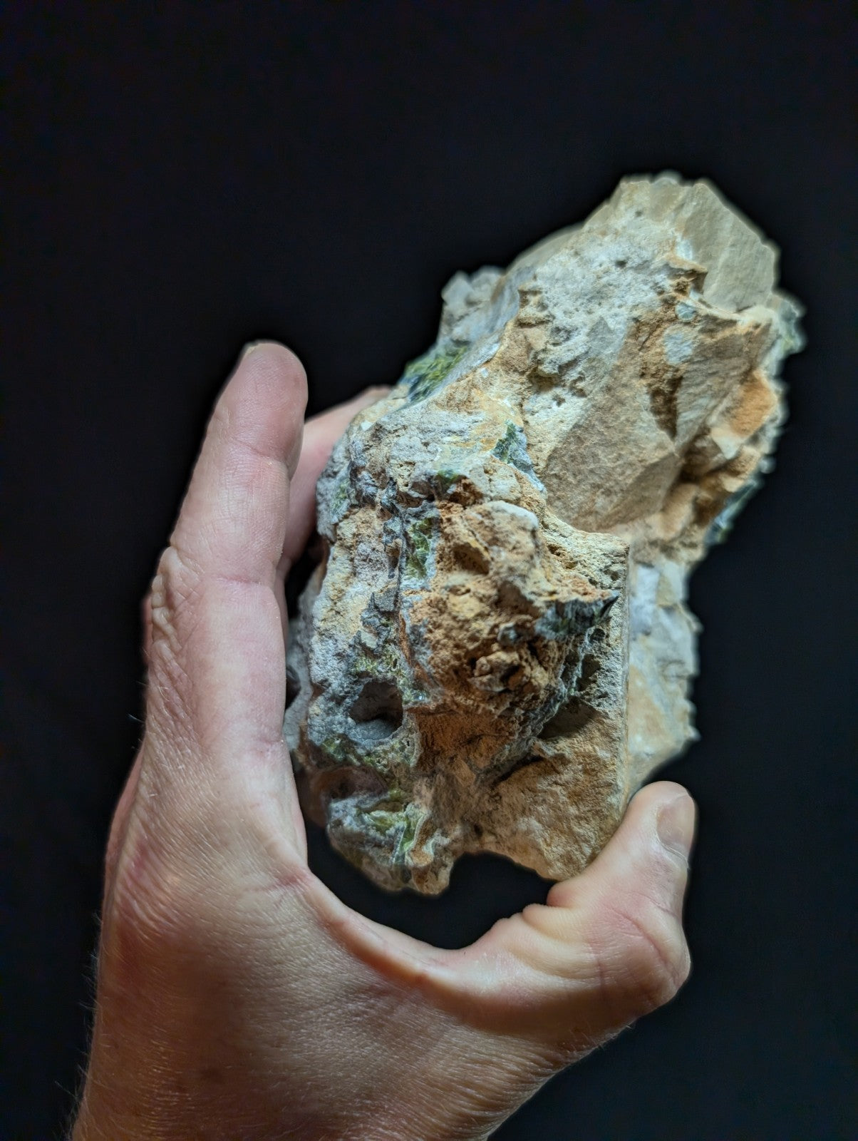 A rugged, irregularly shaped rock with a mix of beige, tan, and greenish hues, featuring rough textures and natural mineral deposits.