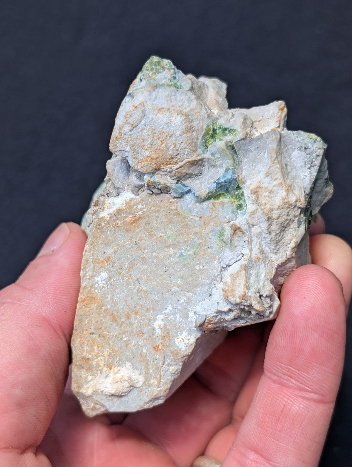 A rough, irregularly shaped mineral specimen with a predominantly grayish-white surface accented by patches of green and subtle orange hues.