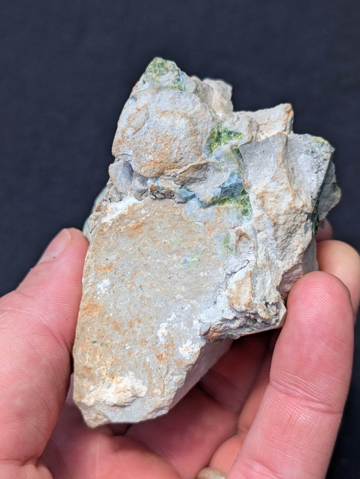 A rough, irregularly shaped mineral specimen with a predominantly grayish-white surface accented by patches of green and subtle orange hues.