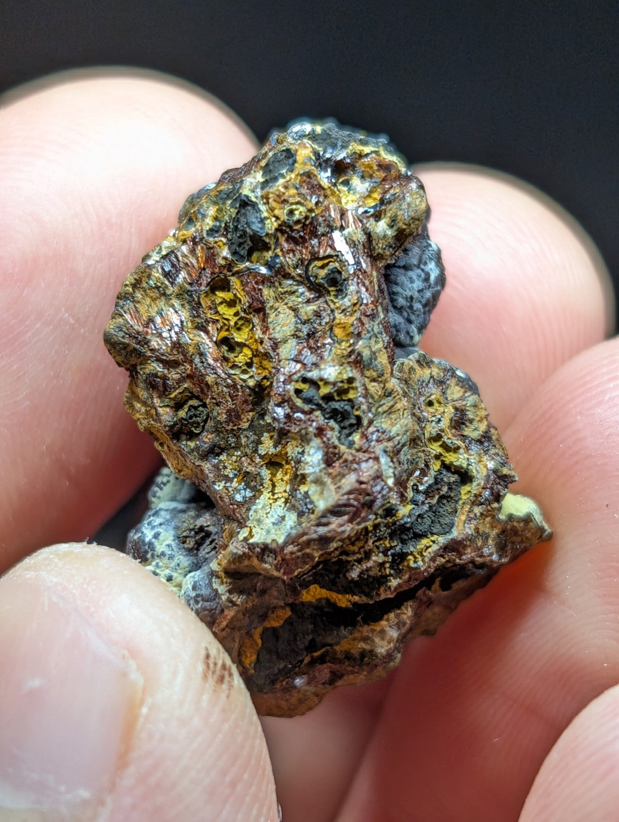 A rugged, irregularly shaped mineral specimen with a mottled surface of dark brown, yellowish-green, and bluish-gray patches.