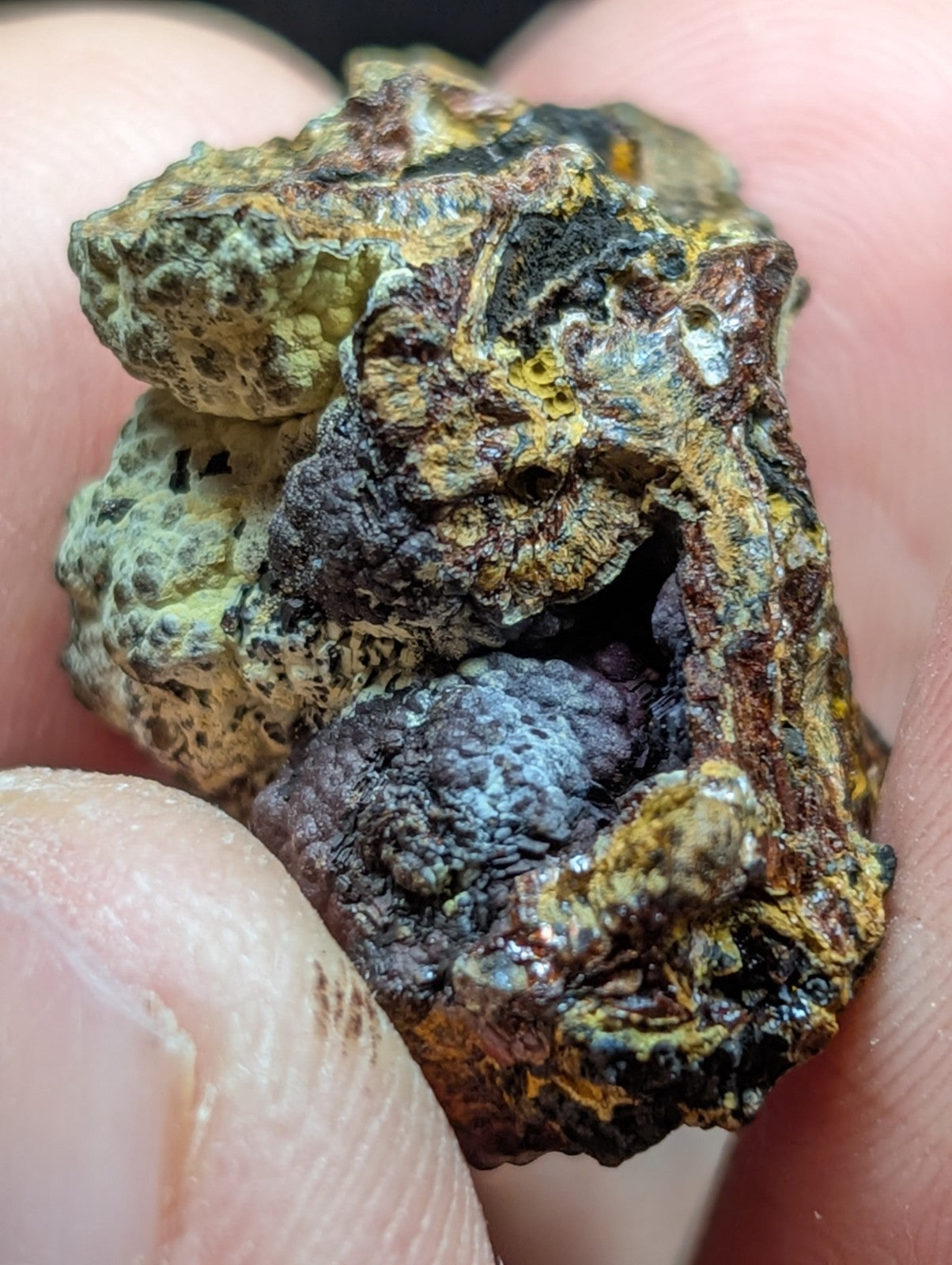 A rugged, multi-colored mineral specimen with patches of yellow, green, black, and hints of purple.