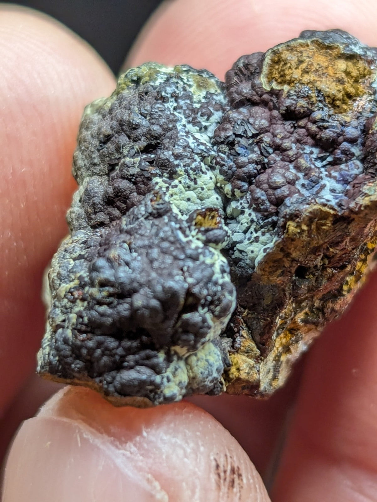 A rugged, irregularly shaped mineral specimen with a textured surface featuring patches of dark gray, greenish-blue patina and areas of yellow-orange oxidation.