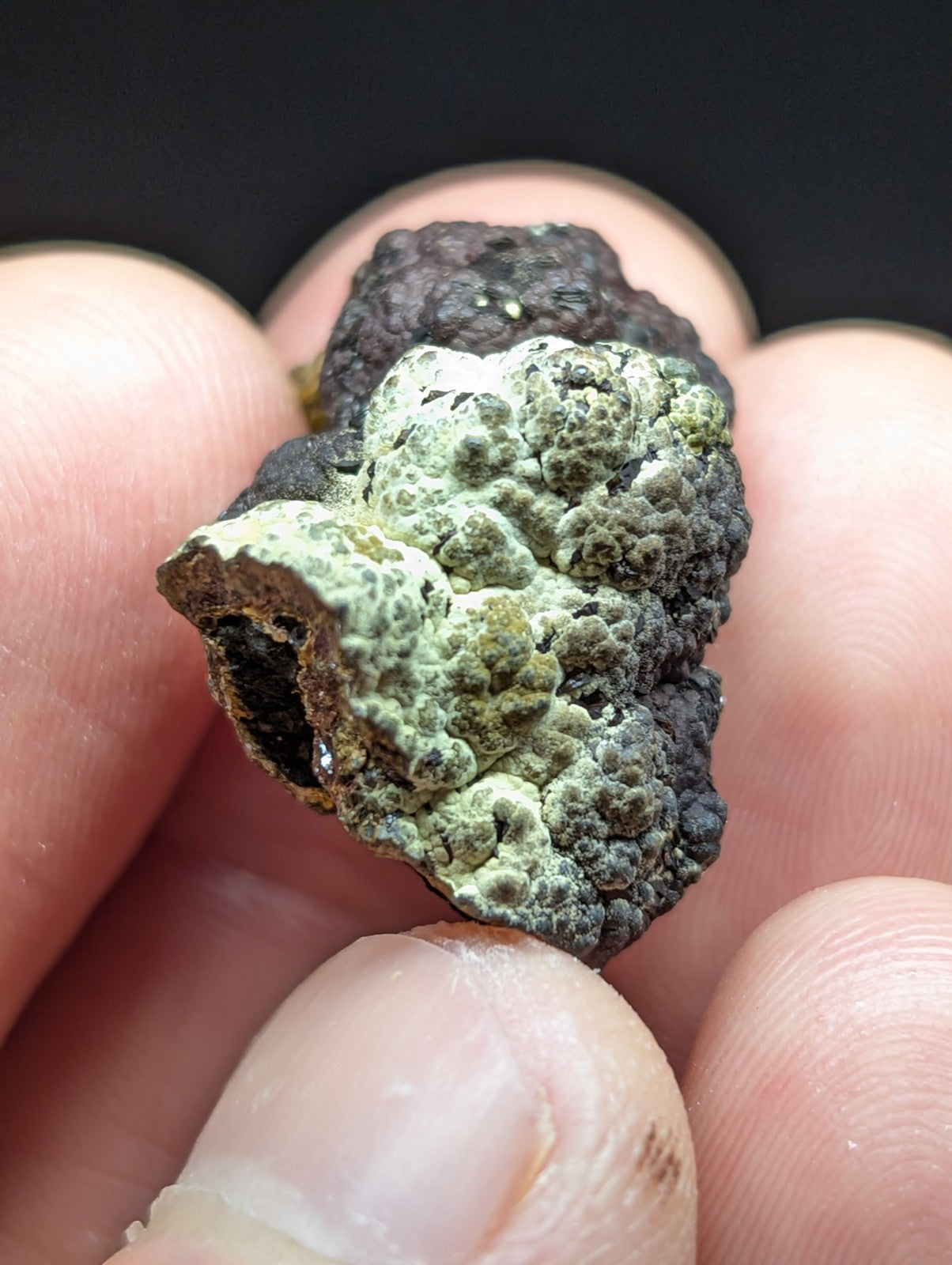 A rough, irregularly shaped mineral specimen with a textured surface featuring patches of greenish lichen-like growth over dark, rocky material.