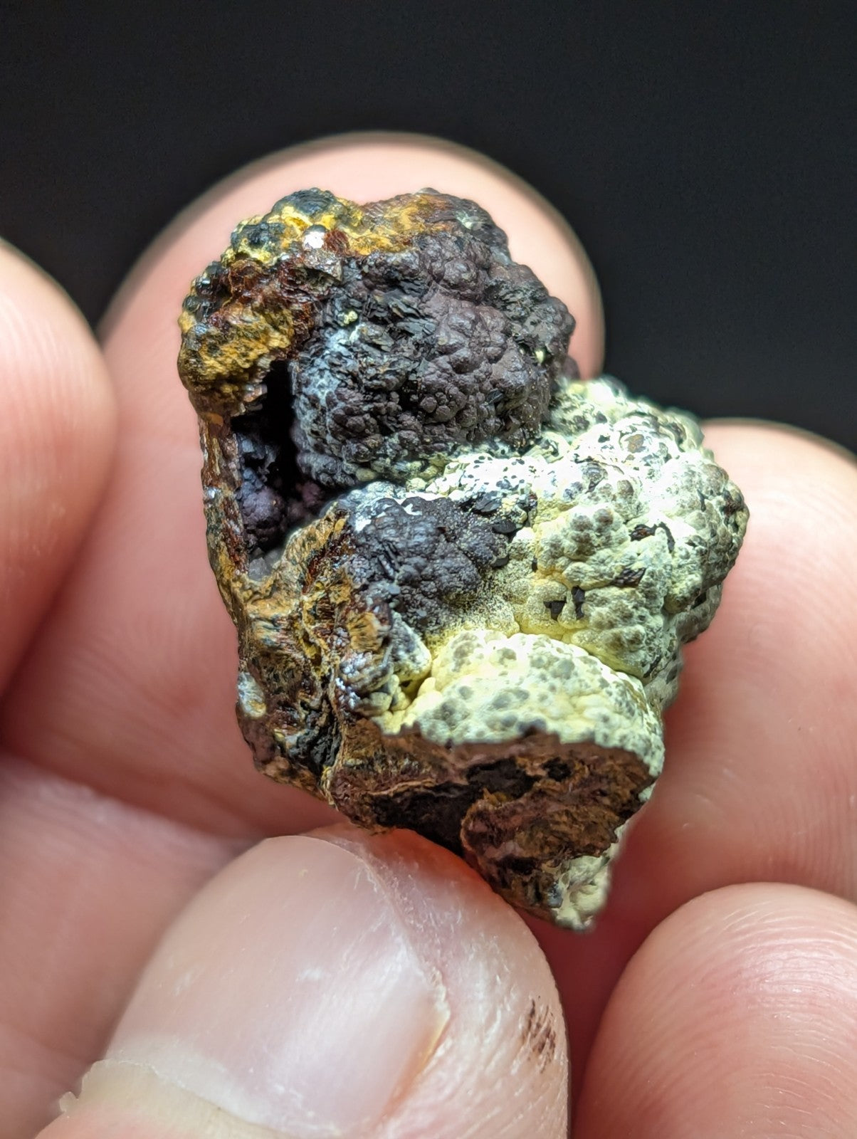 A rugged, multi-colored mineral specimen with patches of greenish-yellow and dark gray/black textures.