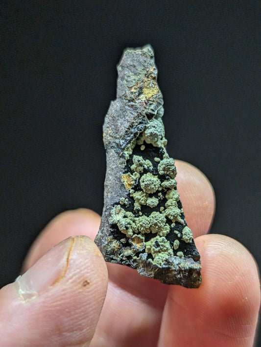 A weathered, irregularly shaped fragment of metal with patches of green patina and small yellowish mineral deposits.