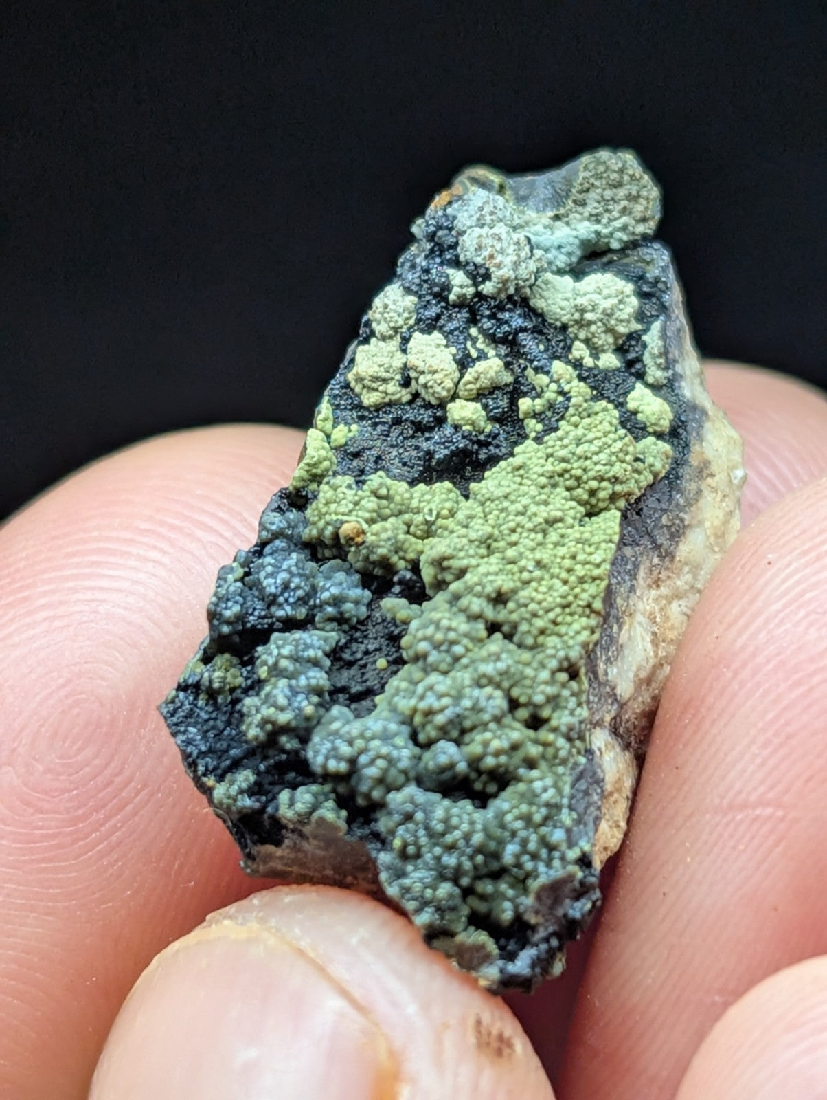 A textured, irregularly shaped mineral specimen featuring patches of vibrant green lichen-like growth over a dark, rough surface.