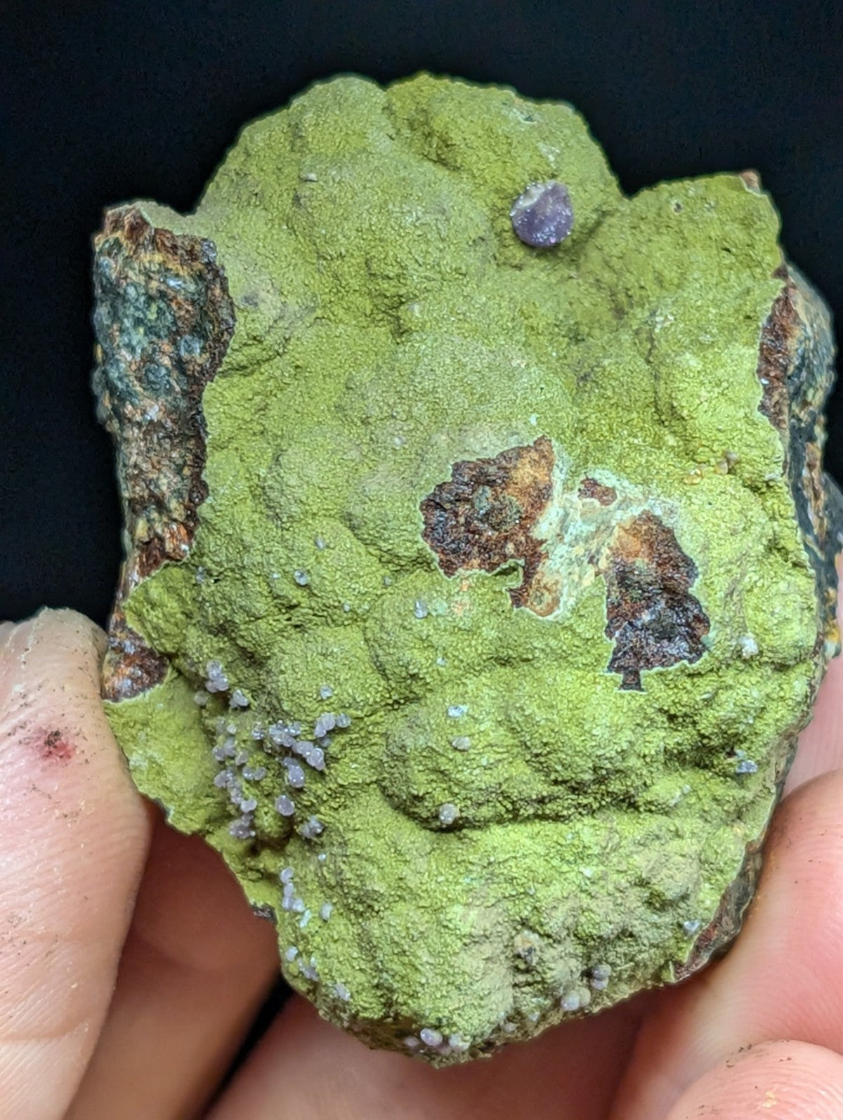 A rough, irregularly shaped green mineral specimen with patches of brown and purple inclusions.