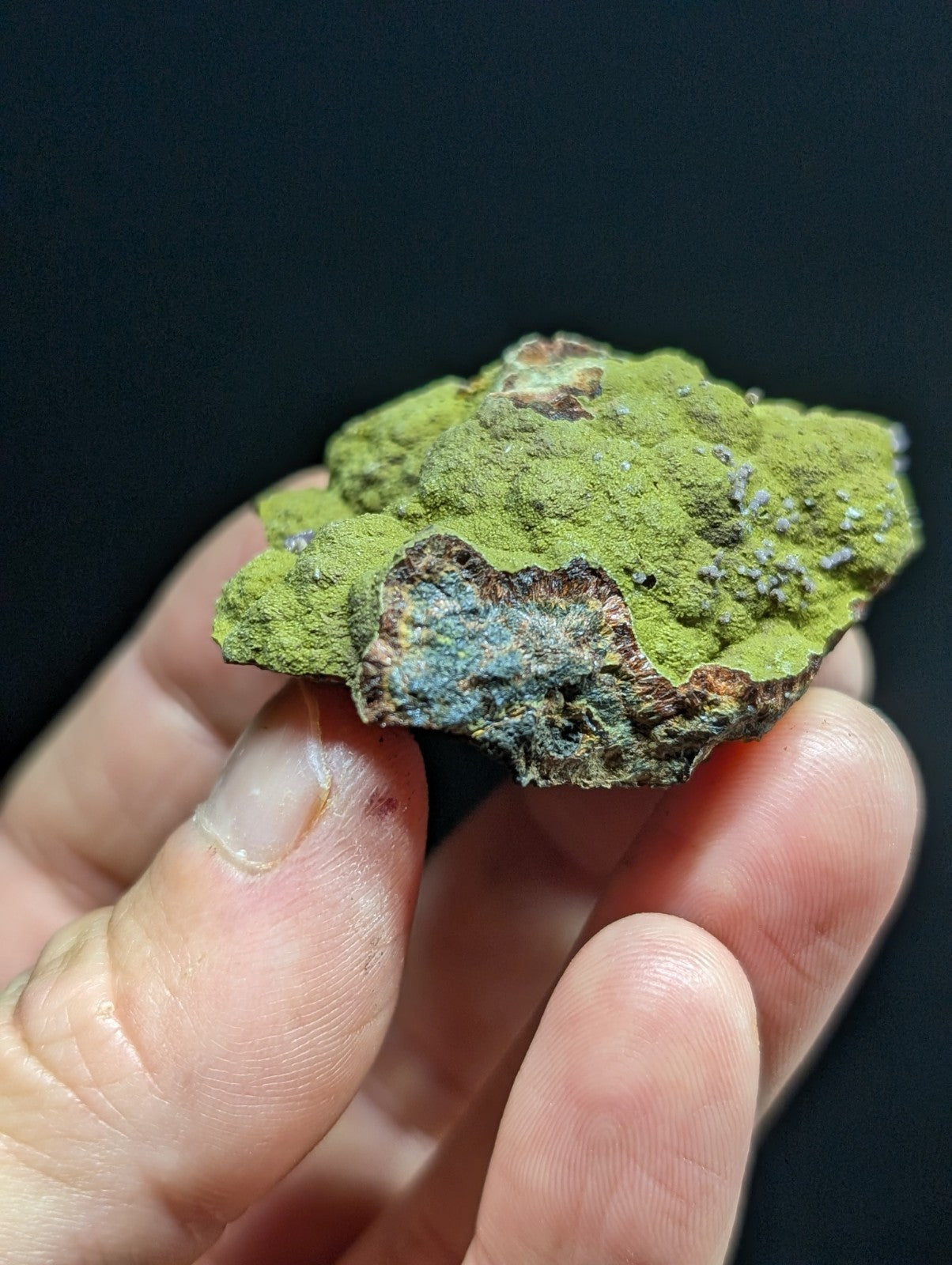A hand holds a rough, irregularly shaped mineral specimen with vibrant green moss-like coating over exposed brown and blue-gray rock.