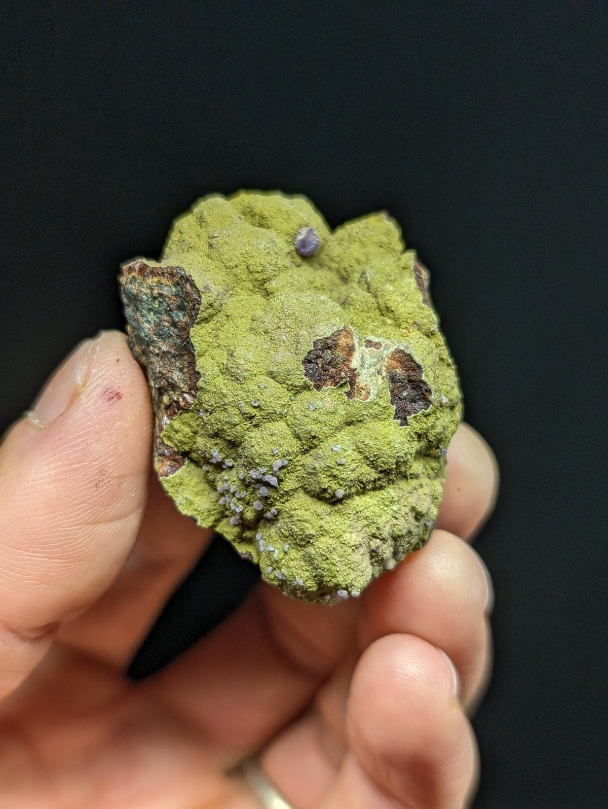 A hand holds a rough, irregularly shaped mineral specimen with vibrant green moss-like coating and scattered brownish patches.