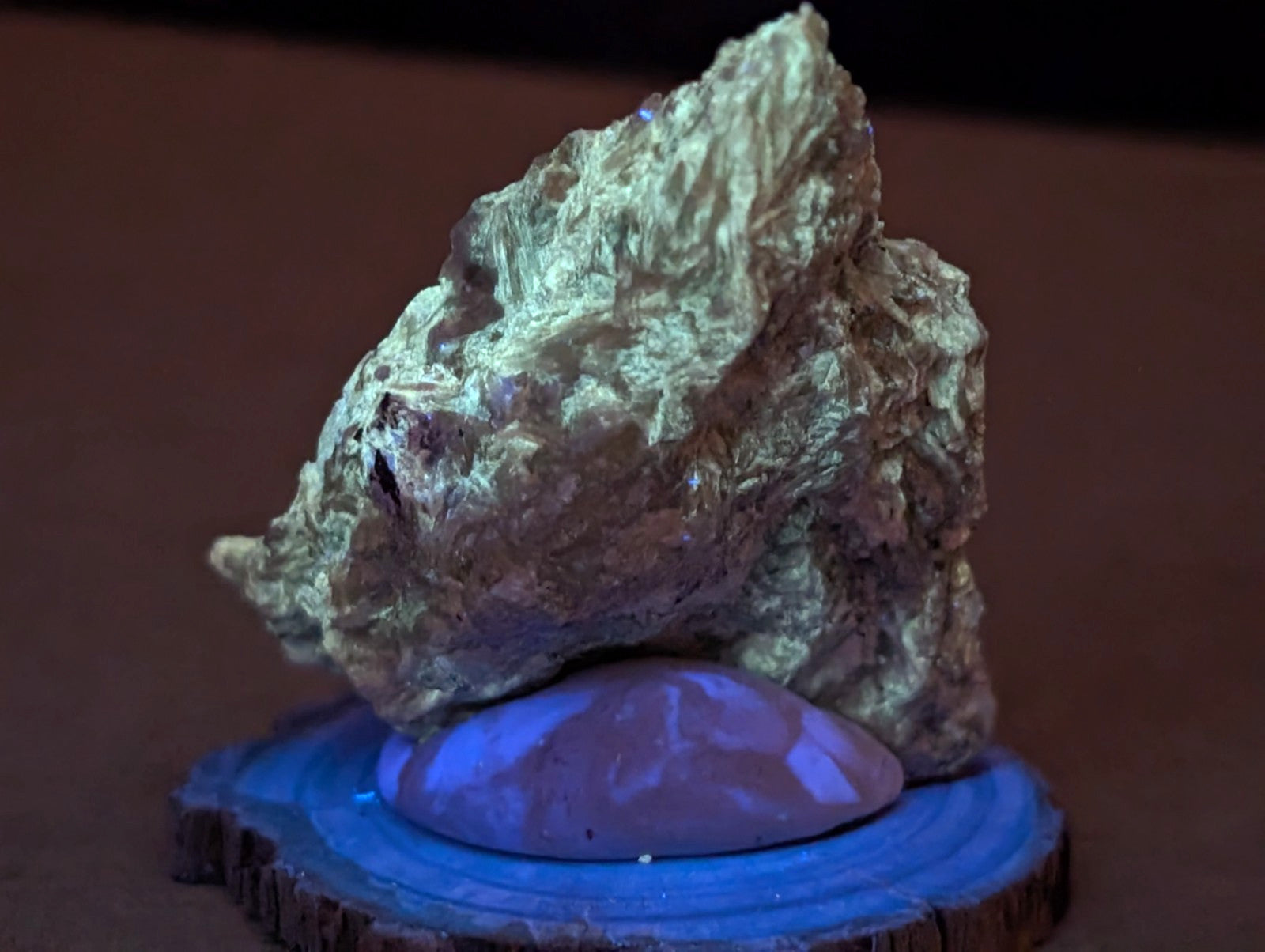 A textured, irregularly shaped gold-colored mineral specimen with a rough surface and subtle greenish undertones rests on a circular wooden base.