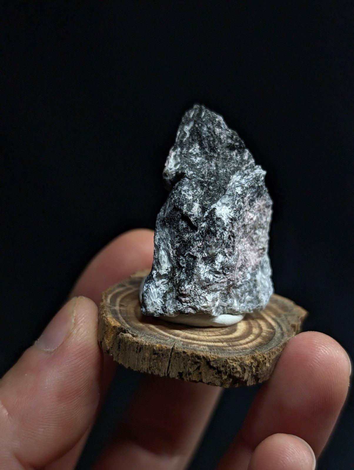 A dark, rough-textured mineral specimen with speckles of white and subtle pink hues rests on a circular wooden slice.