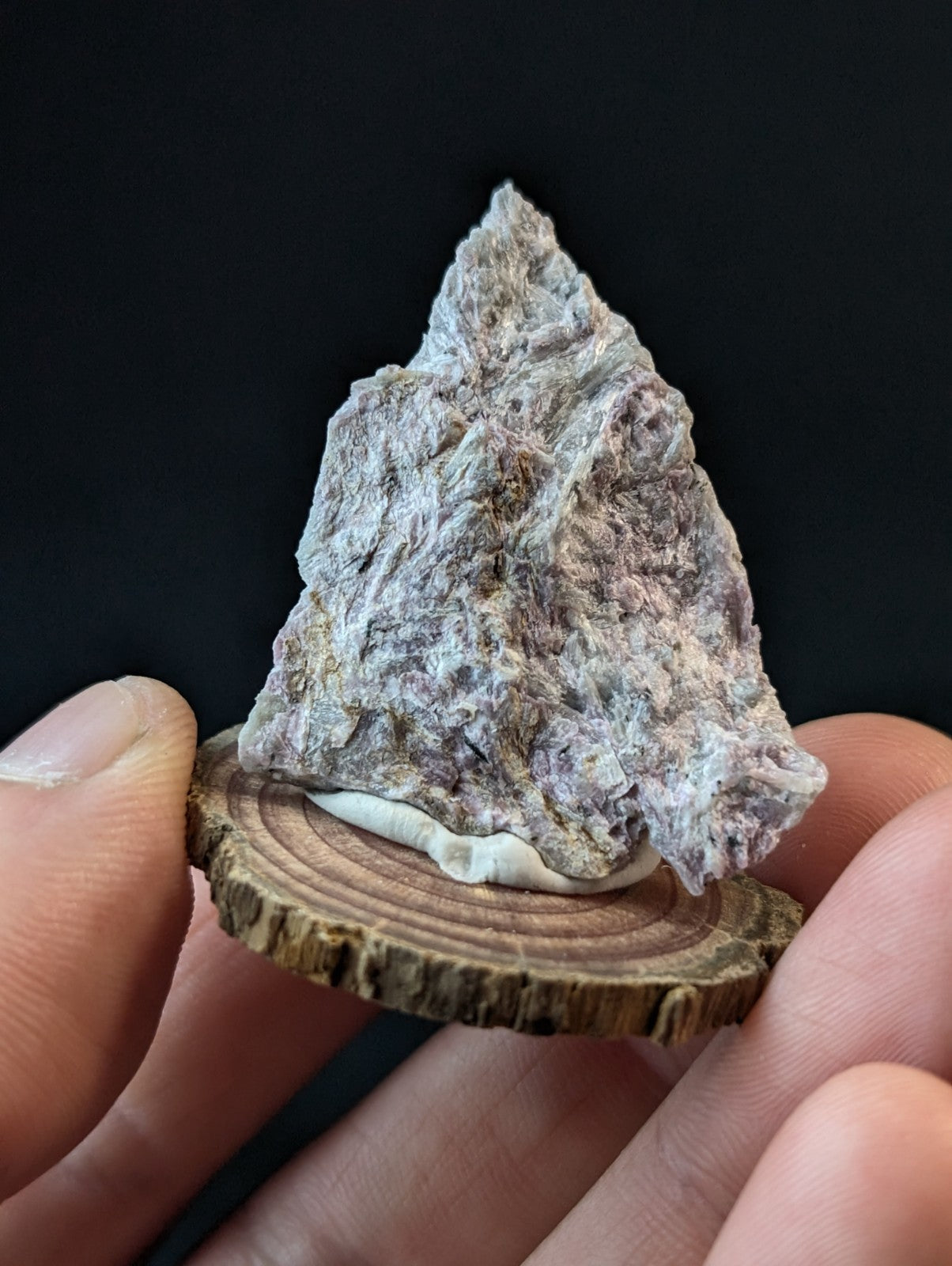 A textured, grayish-purple mineral specimen with a rough, jagged surface and subtle pink undertones rests on a circular wooden slice.