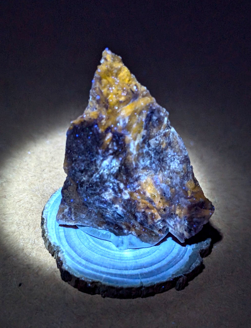 A striking, irregularly shaped mineral specimen with vibrant yellow and deep blue-purple hues rests on a circular wooden slice base.