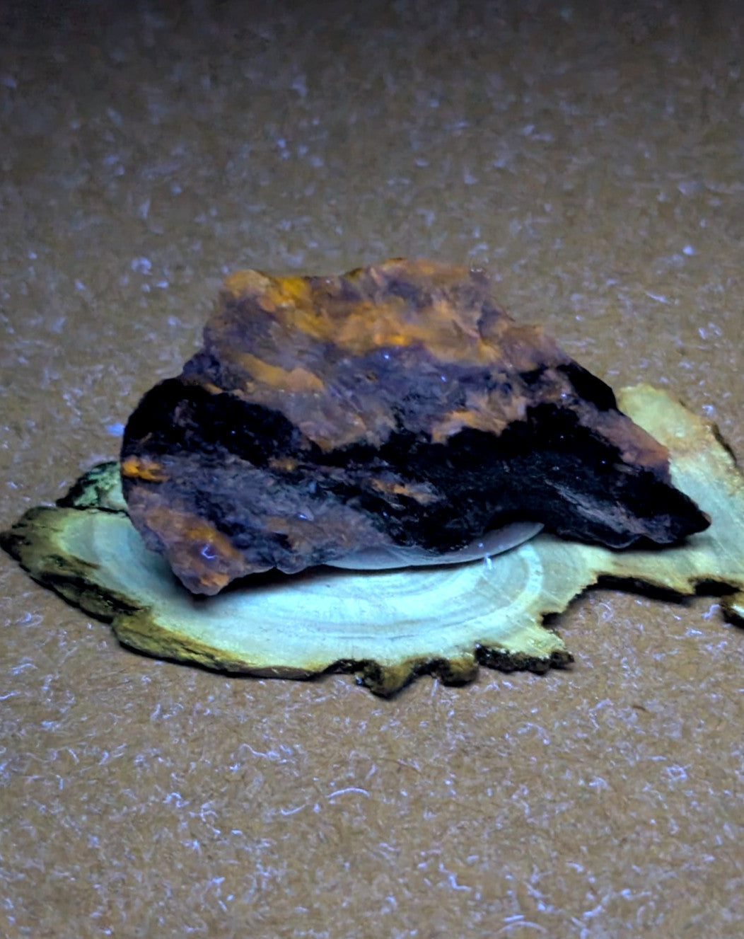 A rugged, irregularly shaped piece of iron ore with a dark metallic base and patches of orange-brown oxidation resting on a light-colored wooden slice.